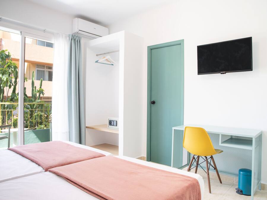 A hotel room with a bed, a desk, a chair, a TV, and a balcony with a view of buildings and plants.