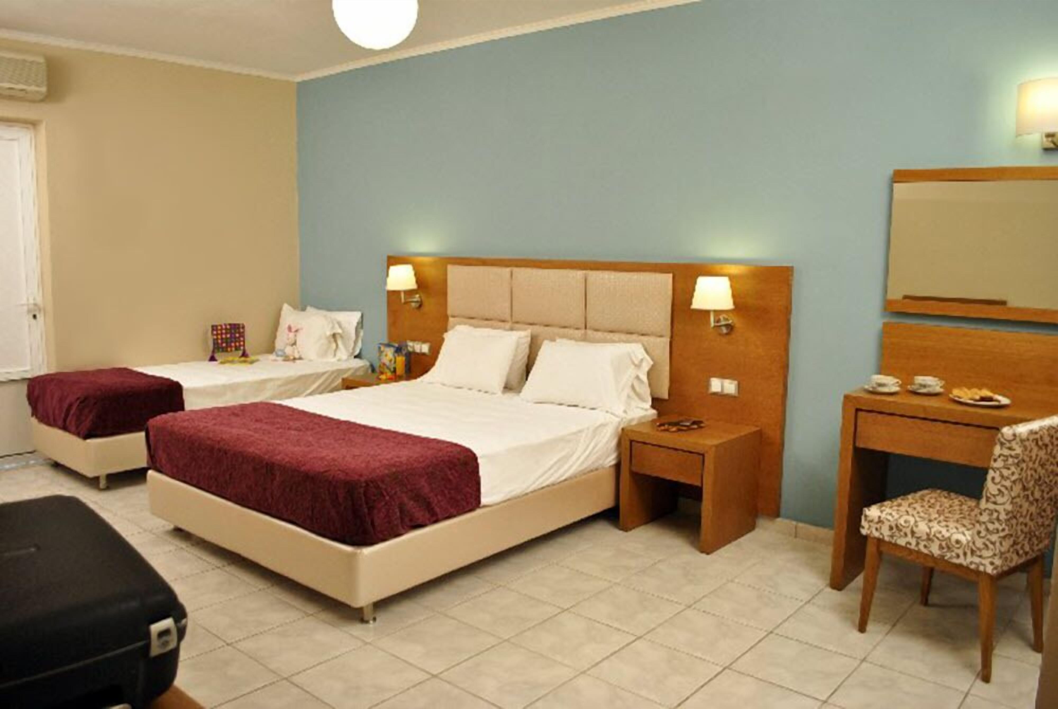 Quarto de hotel com duas camas de solteiro, uma escrivaninha, uma cadeira e uma mesinha de cabeceira com abajur.