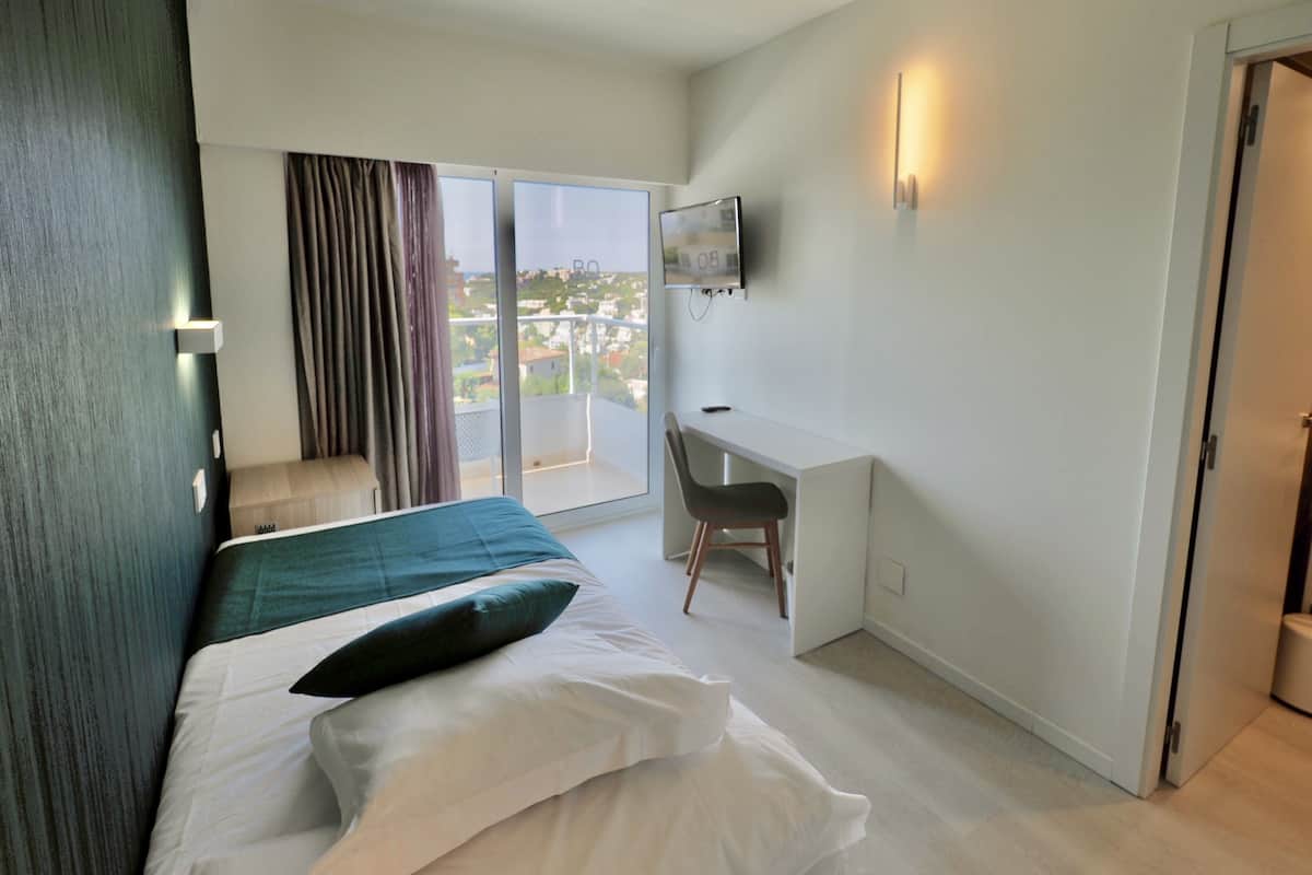 A hotel room with a bed, a desk, a chair, a TV, and a balcony with curtains.