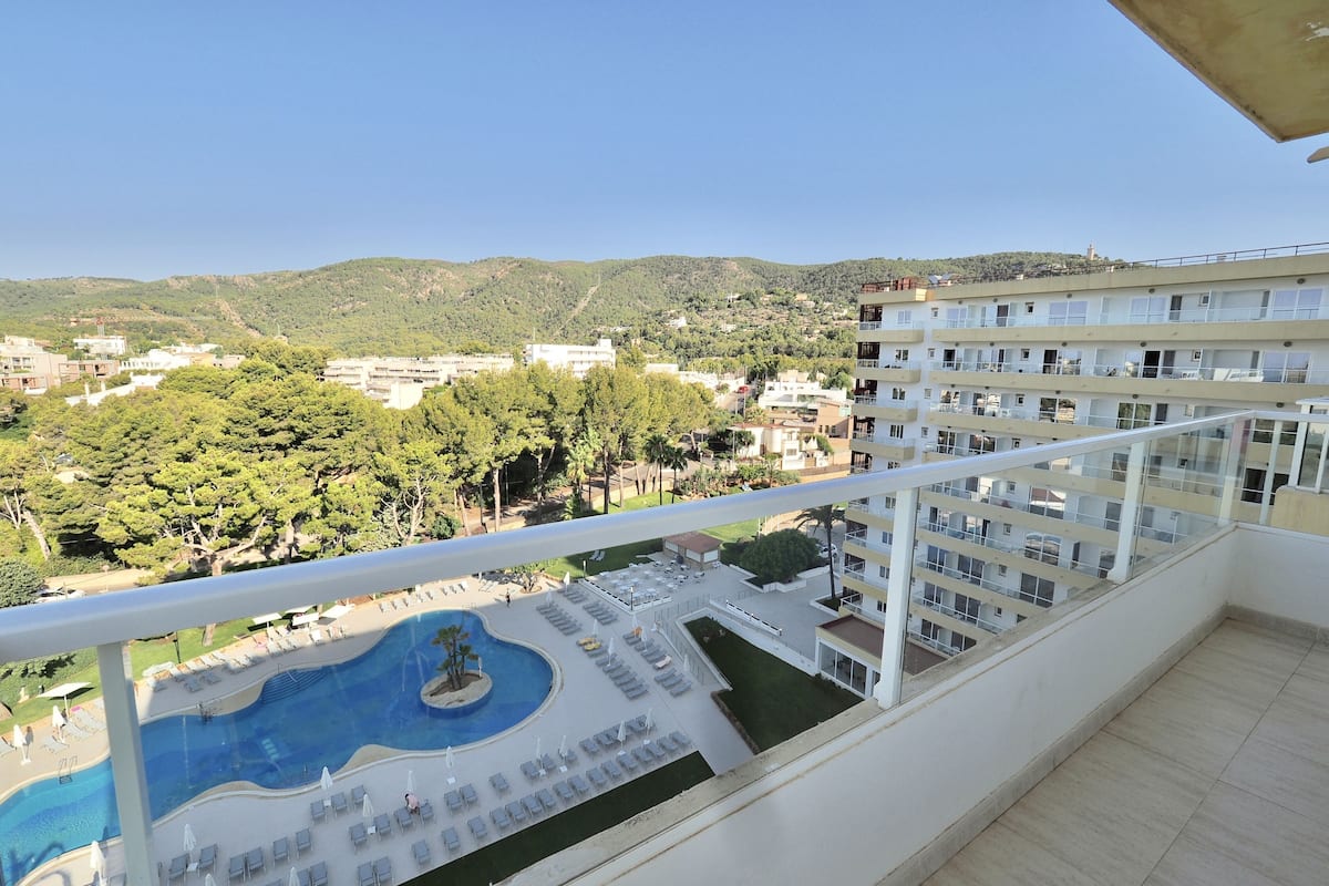 A balcony with a view of a swimming pool and lounge area, surrounded by greenery and buildings.