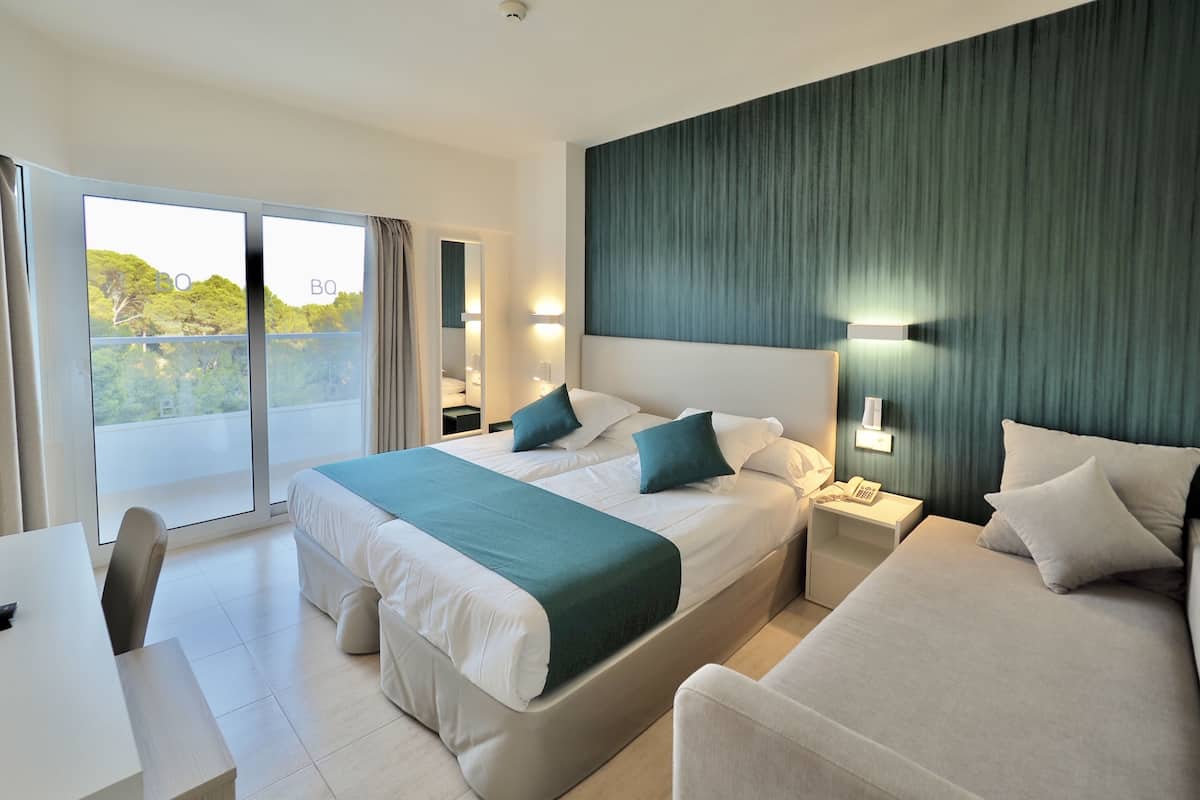 A modern hotel room with a large bed, a sofa, a desk, and a balcony with a view of greenery.