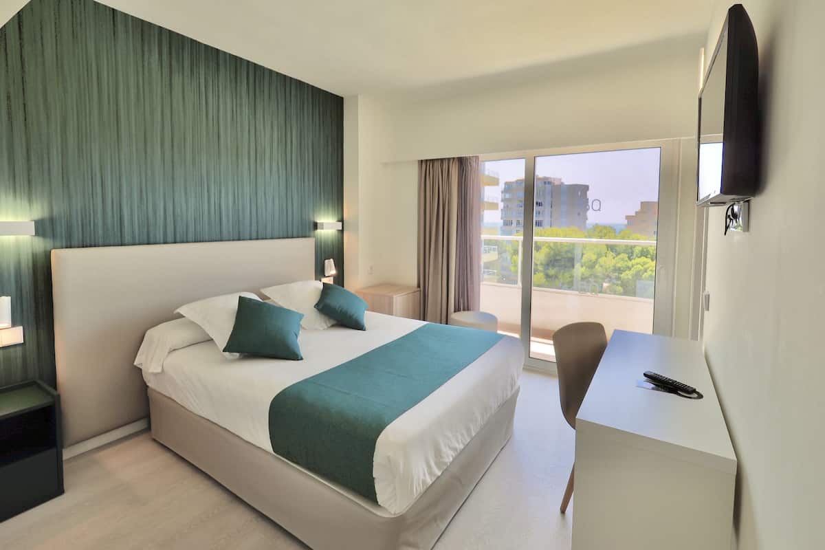 A hotel room with a large bed, a desk, a chair, a TV, and a balcony with a city view.