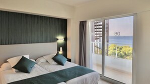 Una habitación de hotel moderna con una cama grande, vista al mar a través de una puerta corrediza de vidrio y un balcón con barandilla.