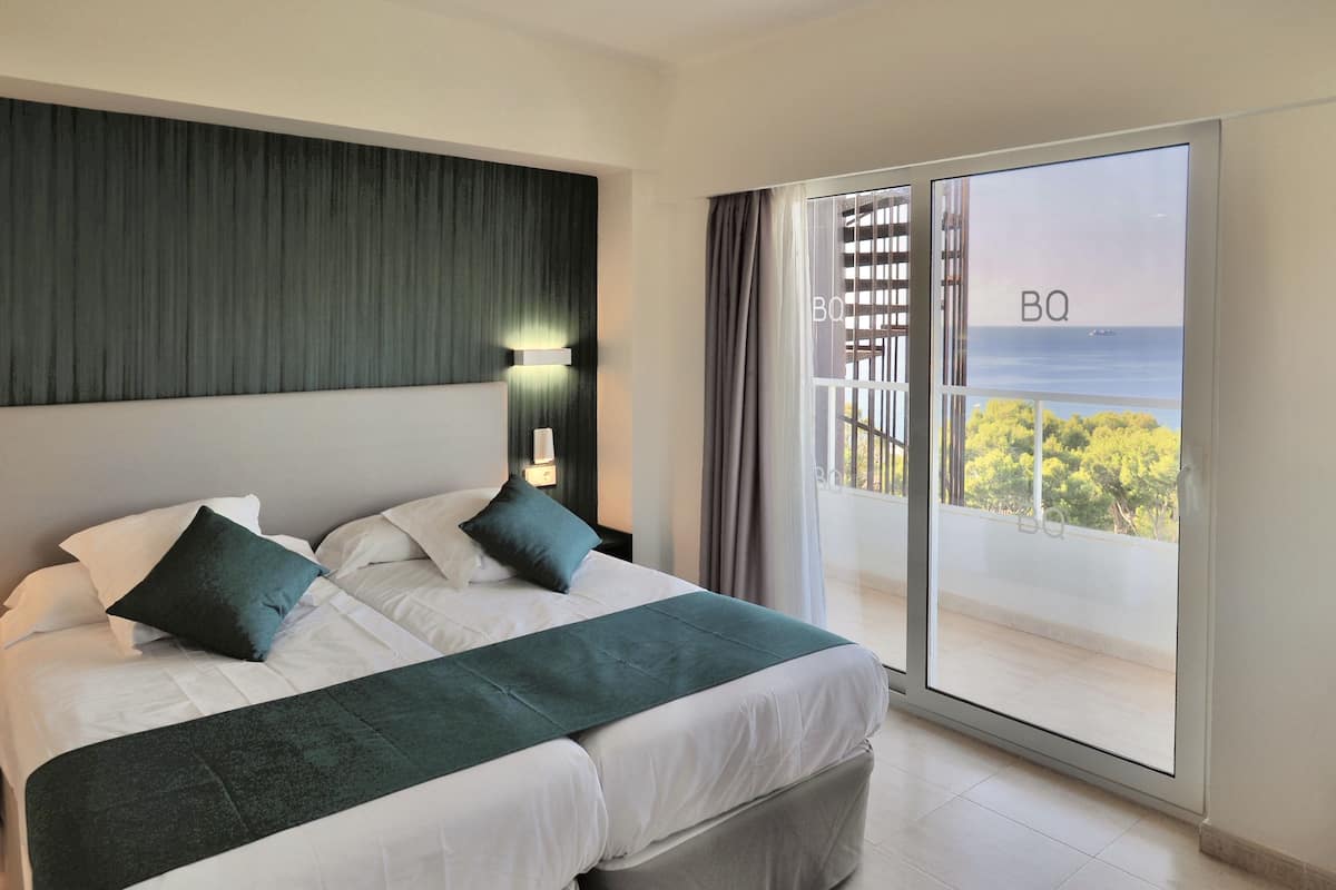 A modern hotel room with a large bed, a view of the sea through a sliding glass door, and a balcony with a railing.