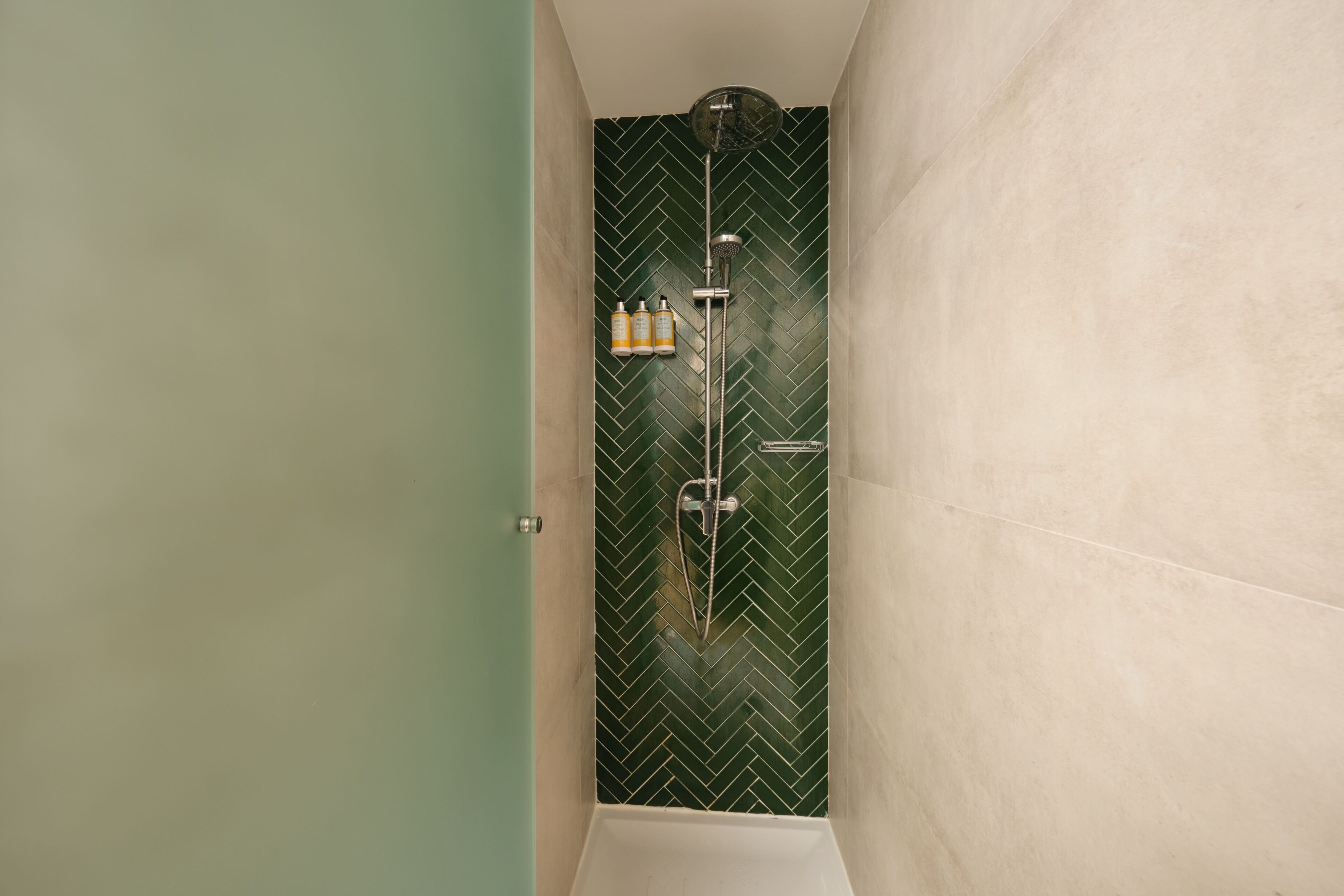 Une salle de bain avec un mur recouvert de carreaux verts disposés en motif chevrons, une douche équipée d’un pommeau de douche à main et une baignoire blanche.