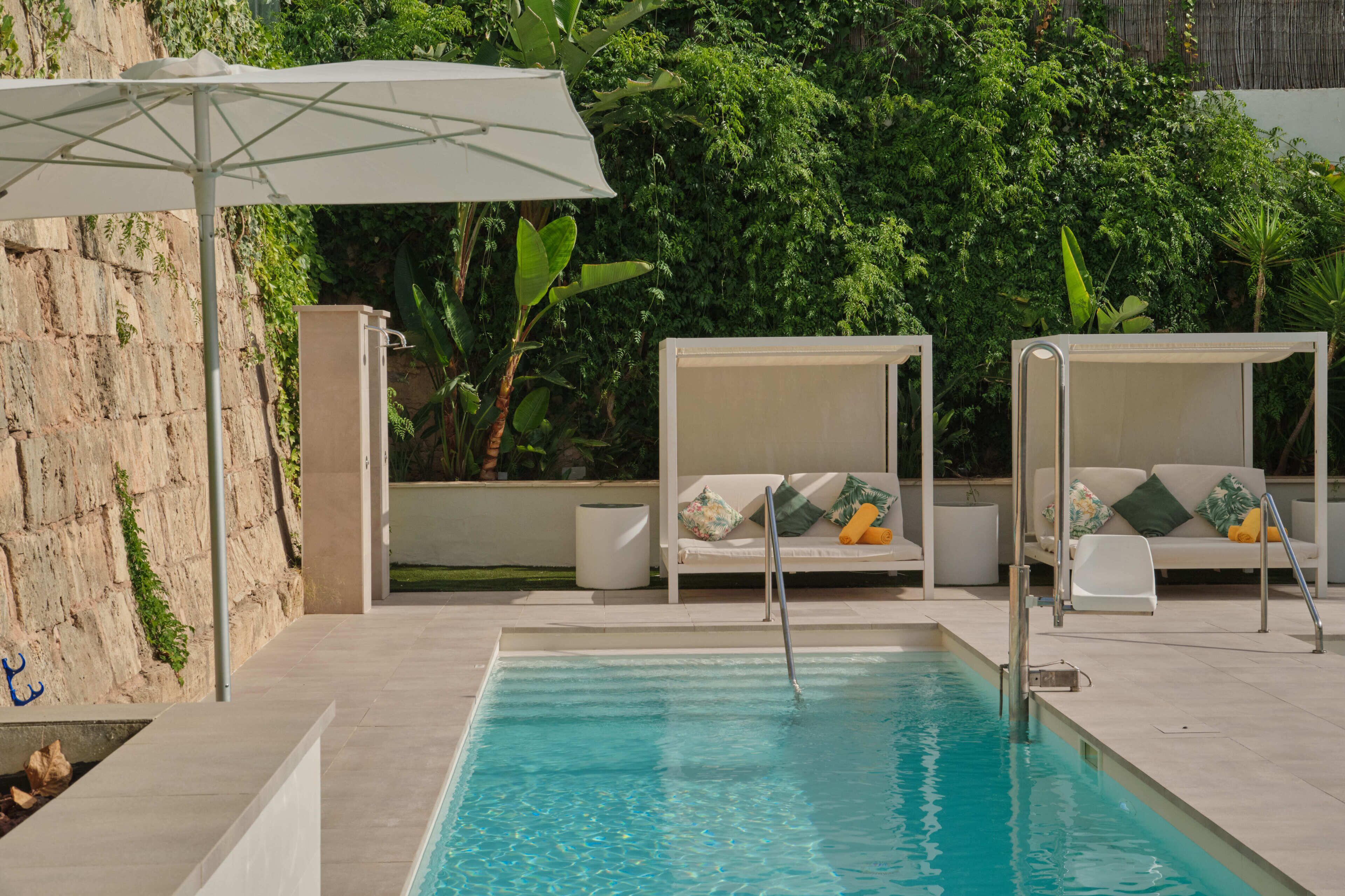 A swimming pool with a white umbrella, two white lounge chairs with cushions, and a stone wall.