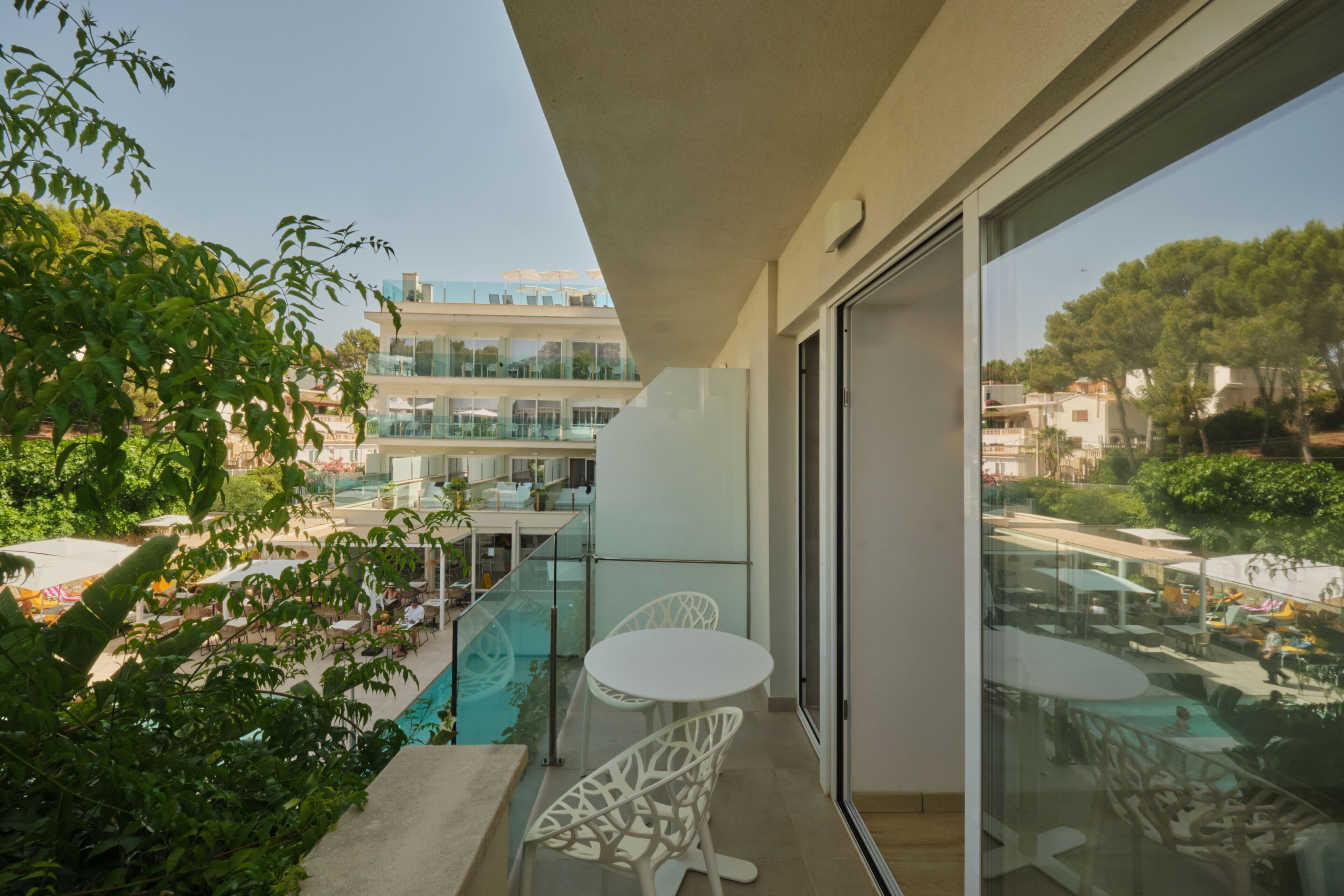 Un balcon avec une porte vitrée, un espace piscine avec des chaises longues et une vue sur un immeuble à plusieurs étages doté d’une terrasse extérieure.
