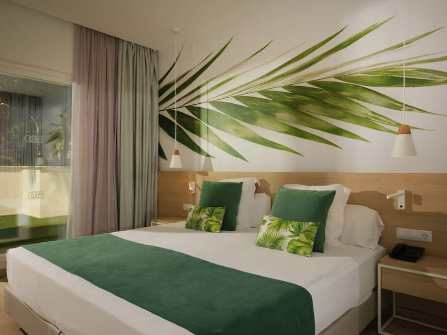 Una habitación de hotel con cama, mesita de noche, lámpara, una planta grande y ventana con cortinas.