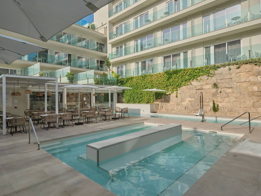 Un espace piscine aménagé avec des chaises longues et des parasols, entouré d’un immeuble à plusieurs étages doté de balcons.