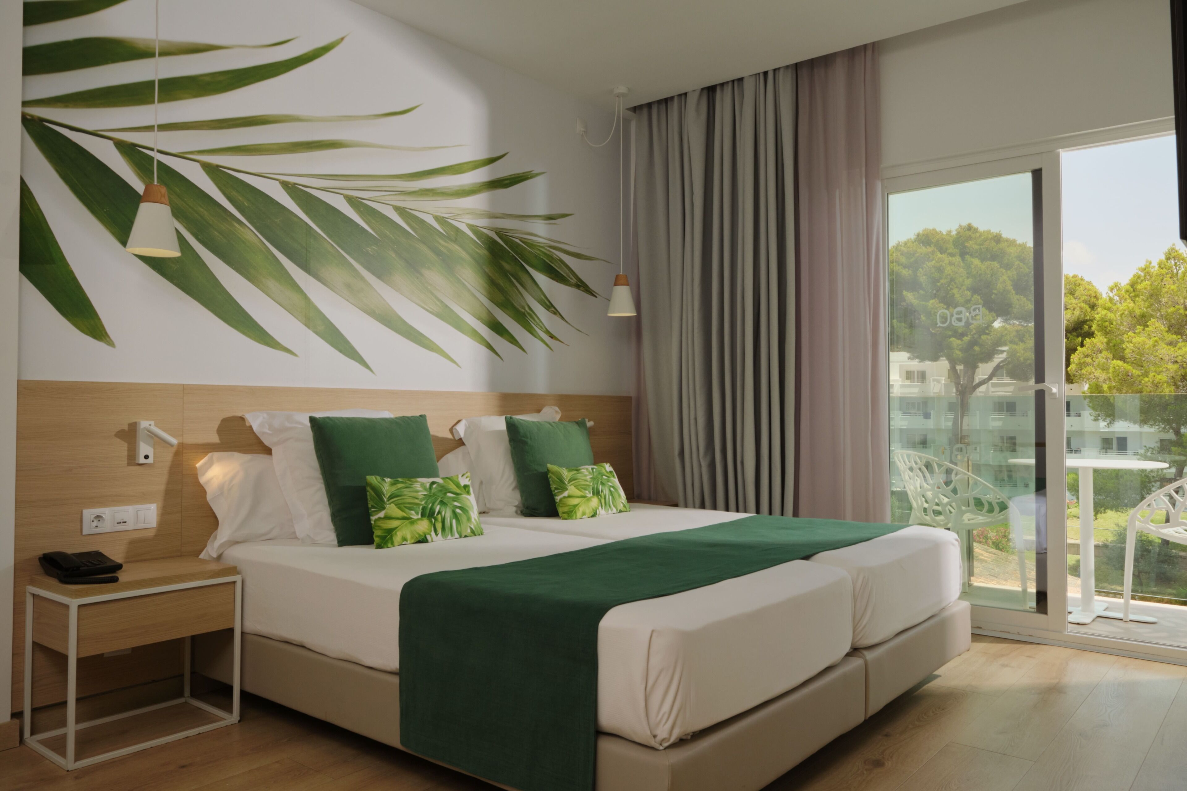 A modern bedroom with a large bed, a nightstand, a lamp, and a view of greenery through the window.