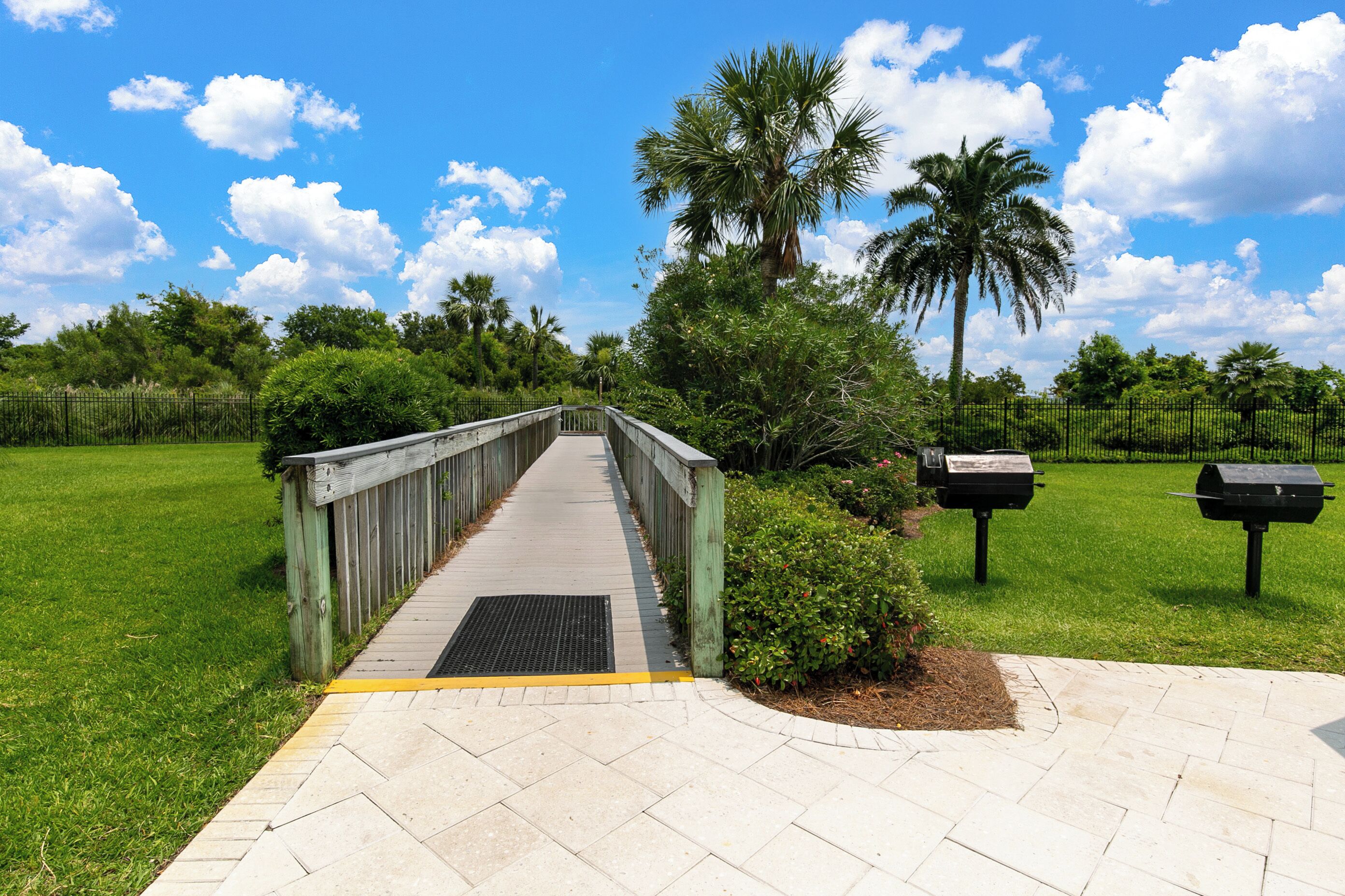 A pathway leading to a wooden bridge over a grassy area with a metal grate, surrounded by lush greenery and palm trees.