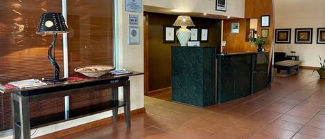 A hotel reception area with a reception desk, a lamp, framed certificates on the wall, and a tiled floor.