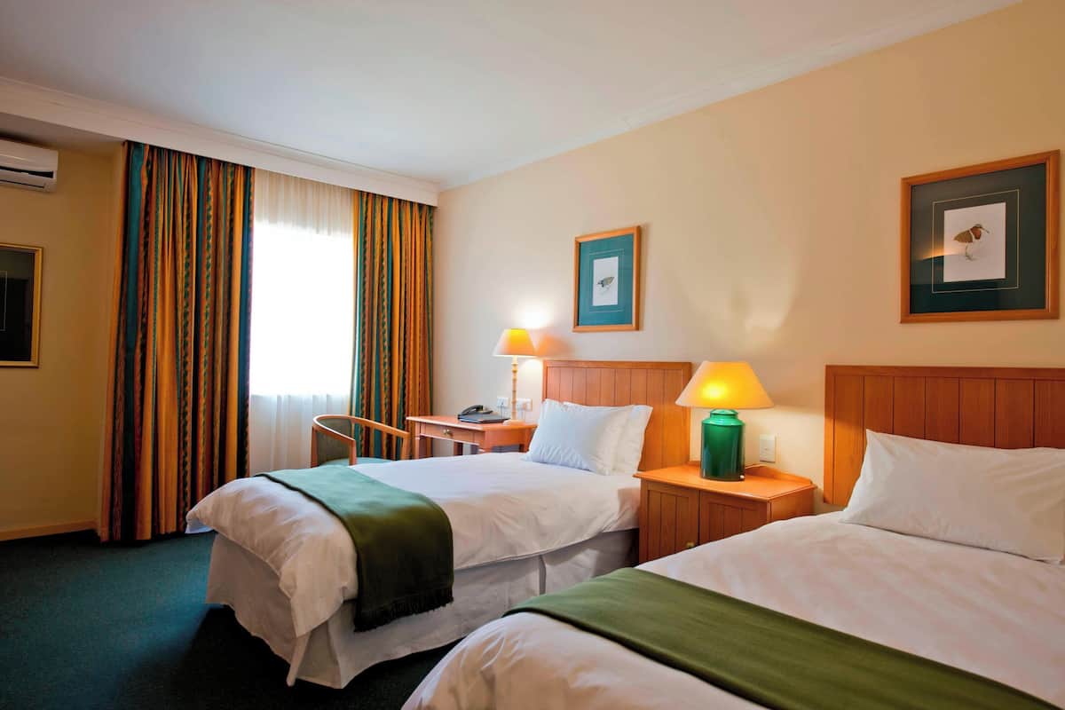 A hotel room with two beds, a desk, a chair, a lamp, and a framed picture on the wall.