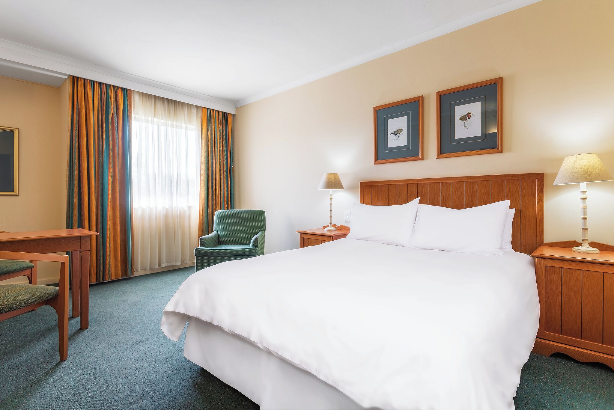Habitación de hotel con cama, escritorio, silla, dos lámparas de noche y dos cuadros en la pared.