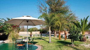 Un espace piscine avec une table et des chaises sous un grand parasol, entouré de palmiers et de verdure.