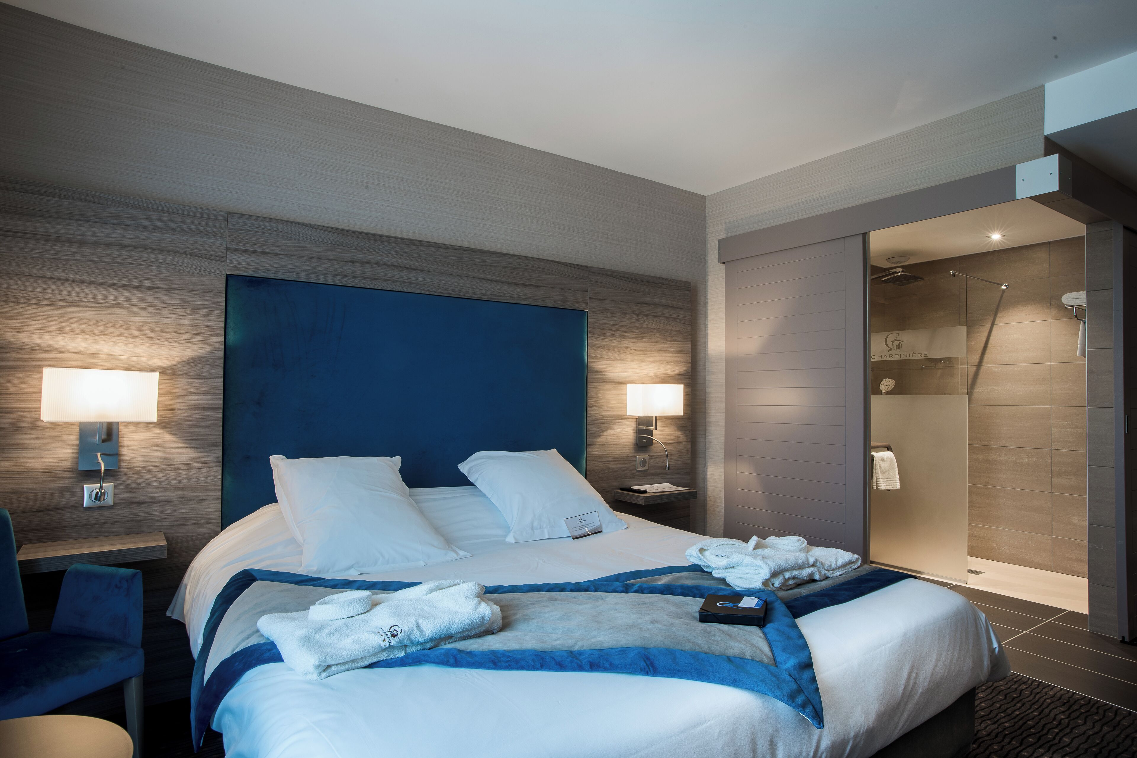Habitación de hotel moderna con una cama grande, dos lámparas de noche, zona de ducha y un área de descanso con una silla.