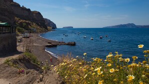 A coastal beach with a rocky cliff, a pier, and boats in the water. - Hotel Mathios Village (Santorini)