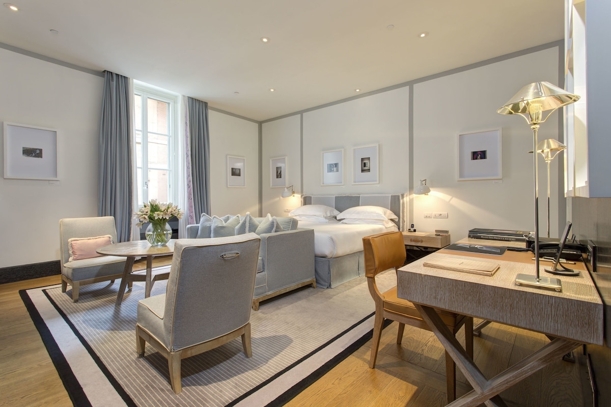 A hotel room with a bed, a desk, two armchairs, a coffee table, and a large window with curtains.