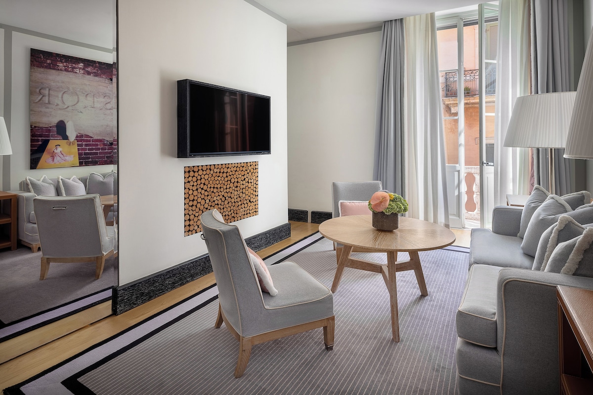 A modern hotel room with a grey sofa, a round wooden table, and a flat-screen TV mounted on the wall.
