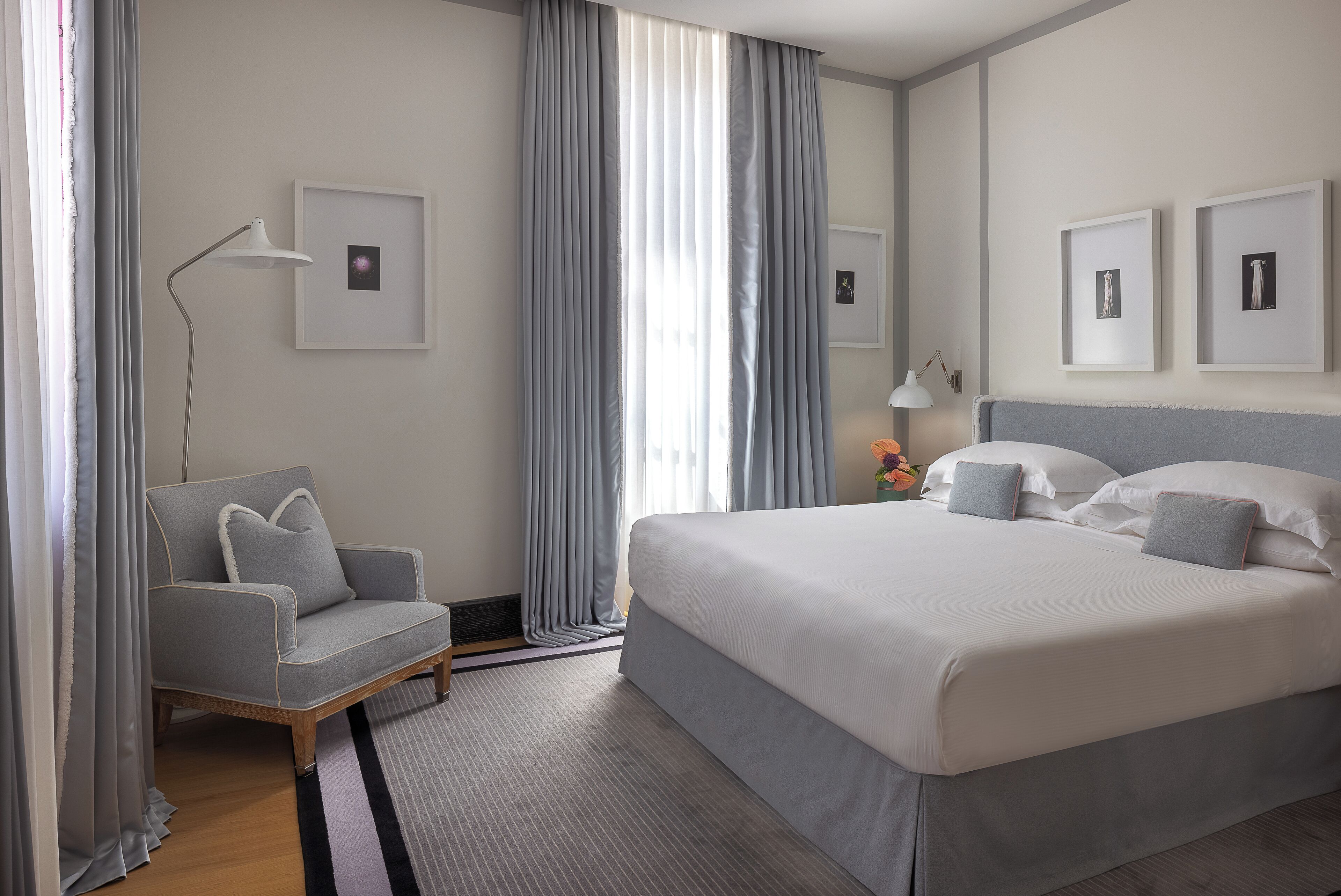 Un cuarto de hotel con una cama grande, un sillón gris, una lámpara de pie y cuadros en la pared.