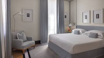 Un cuarto de hotel con una cama grande, un sillón gris, una lámpara de pie y cuadros en la pared.
