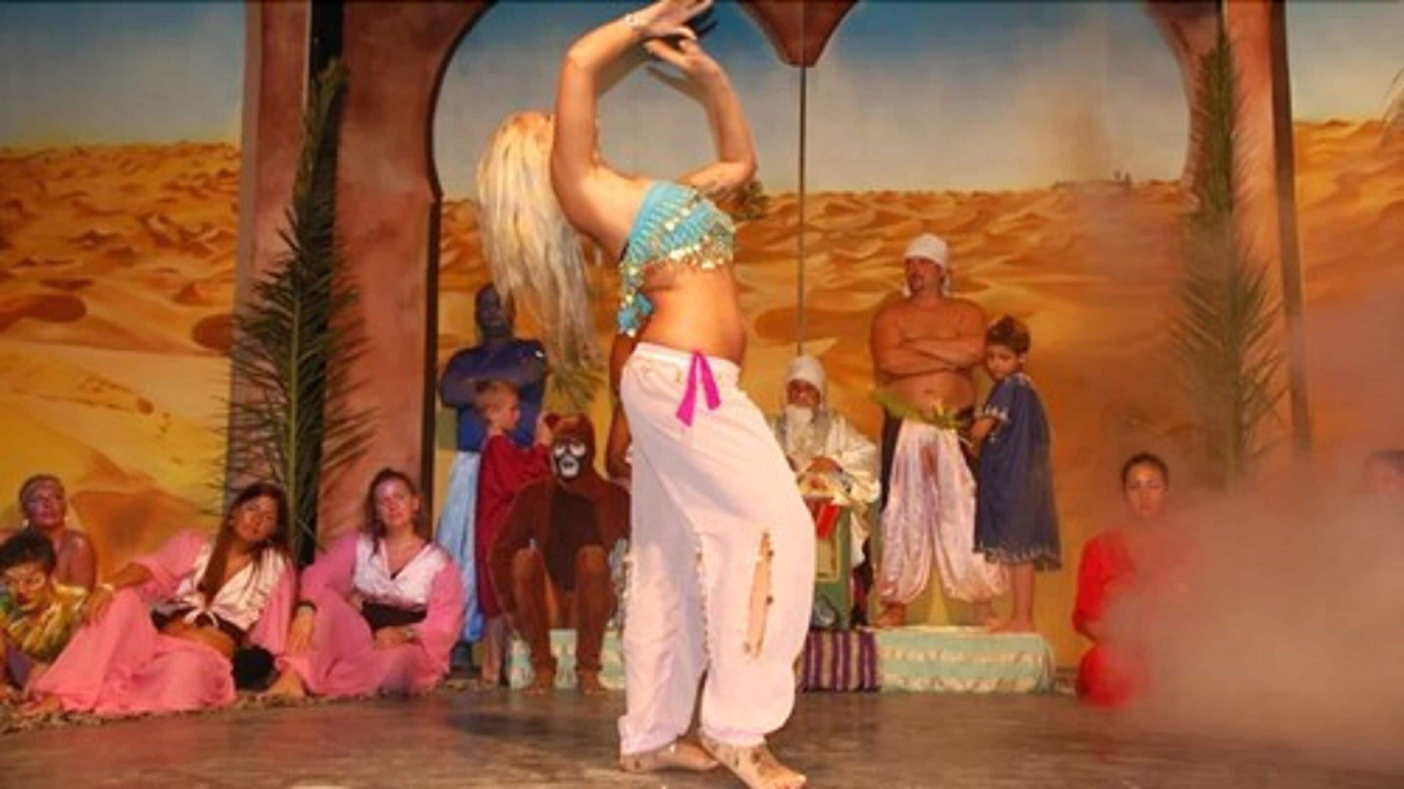 Une scène avec un décor représentant un désert, des artistes vêtus de costumes traditionnels et un public assis par terre.