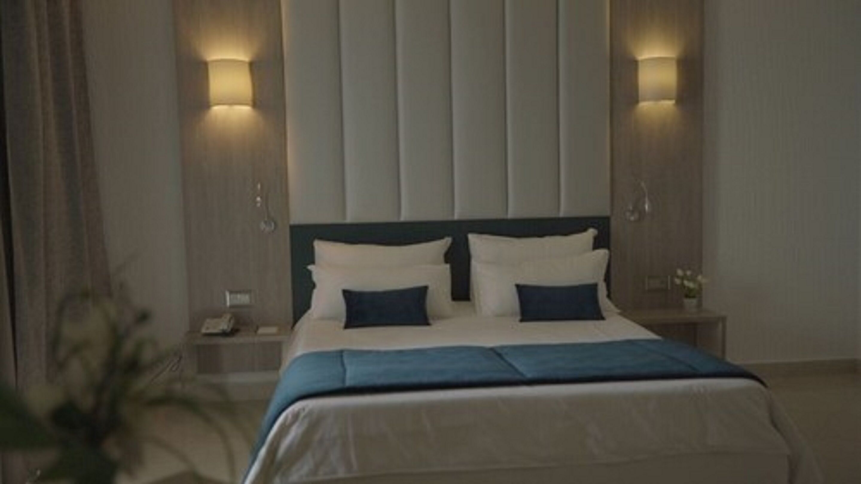 A neatly made bed with a blue accent, two bedside tables with lamps, and a wall with vertical blinds.