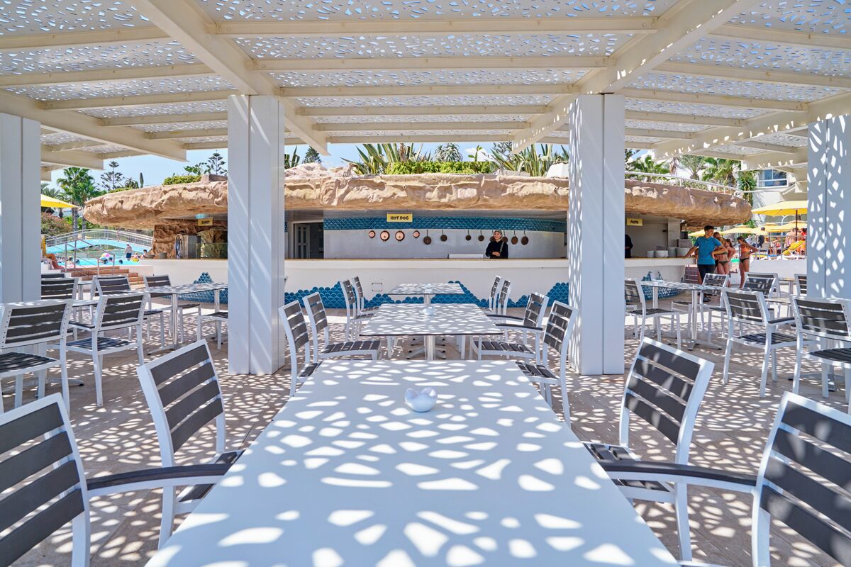 An outdoor dining area with tables and chairs, a rock formation in the background, and a pool area visible to the left.