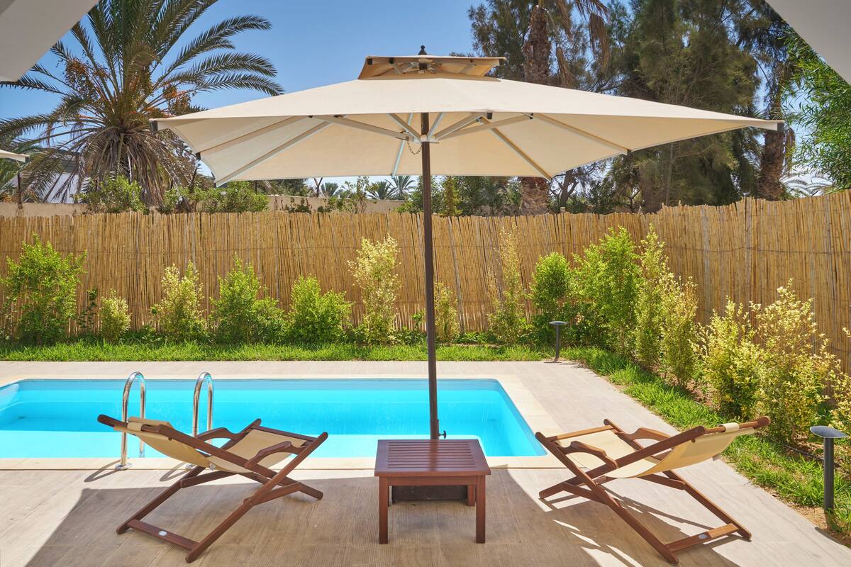 Zona de piscina con dos sillones de descanso y una mesa bajo un sombrilla grande, rodeada por una cerca de bambú y follaje.