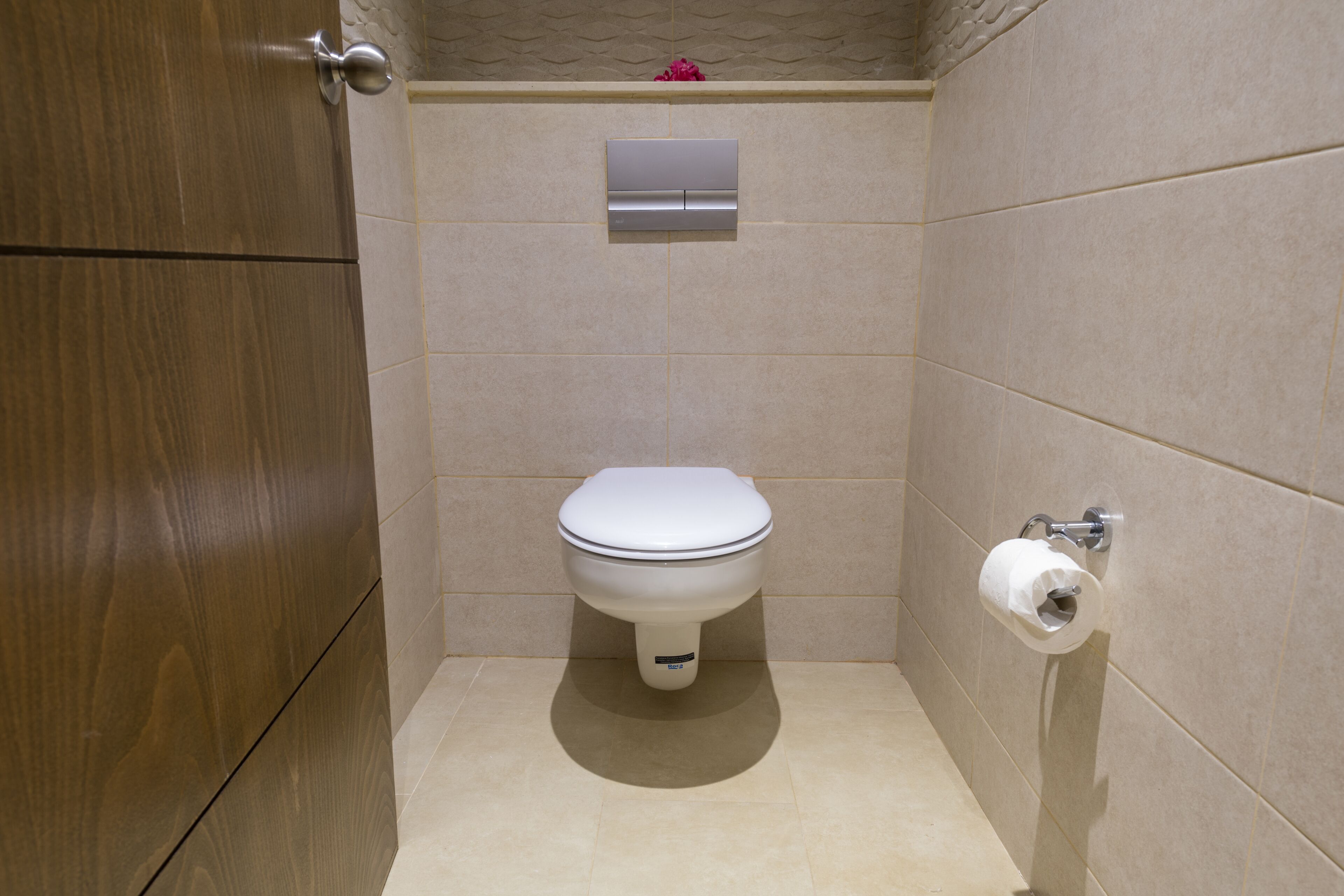 Une petite salle de bain moderne, équipée de toilettes, d’un porte-rouleau de papier toilette fixé au mur et d’une porte en bois.