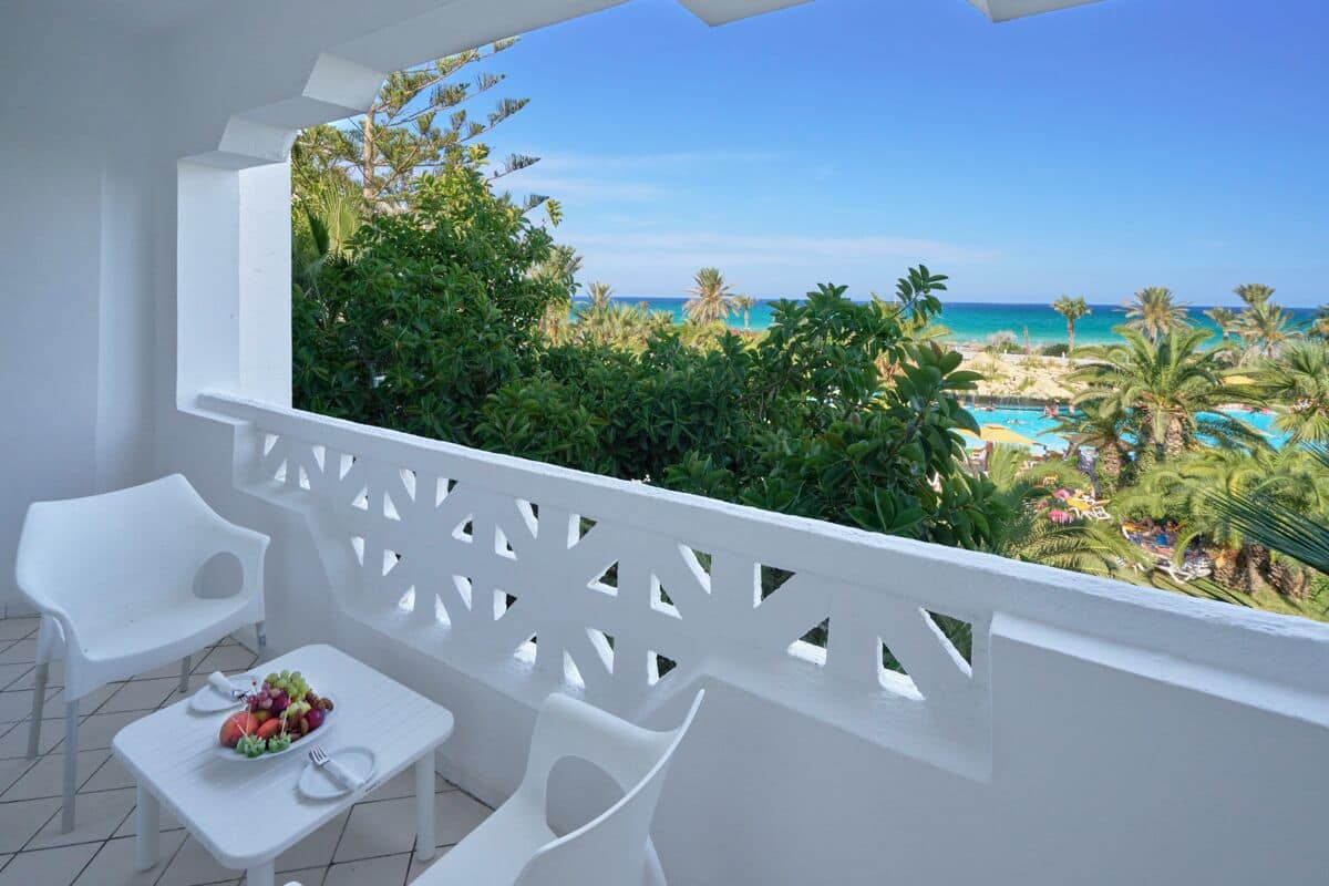 Un balcón con mobiliario blanco, una mesa con frutero y vistas a la playa y la piscina.