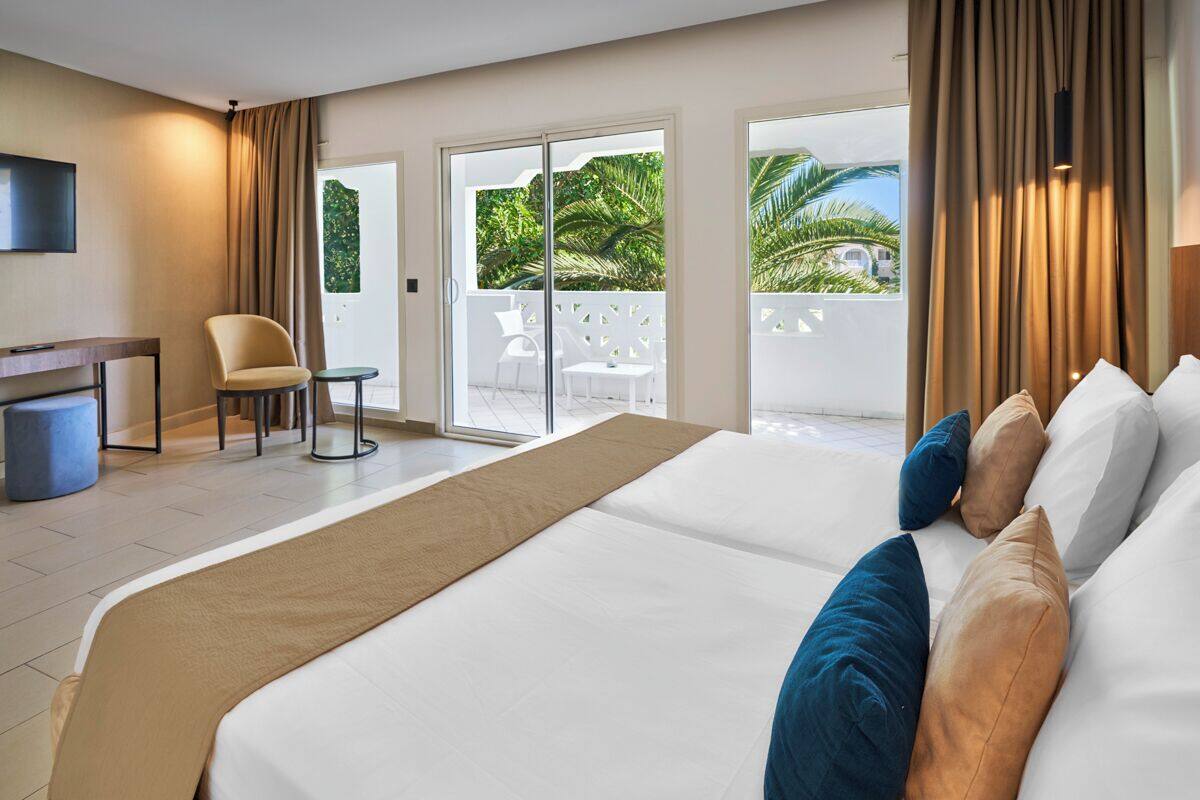 Habitación de hotel con una cama grande, un escritorio, una silla y vistas a un balcón con palmeras.