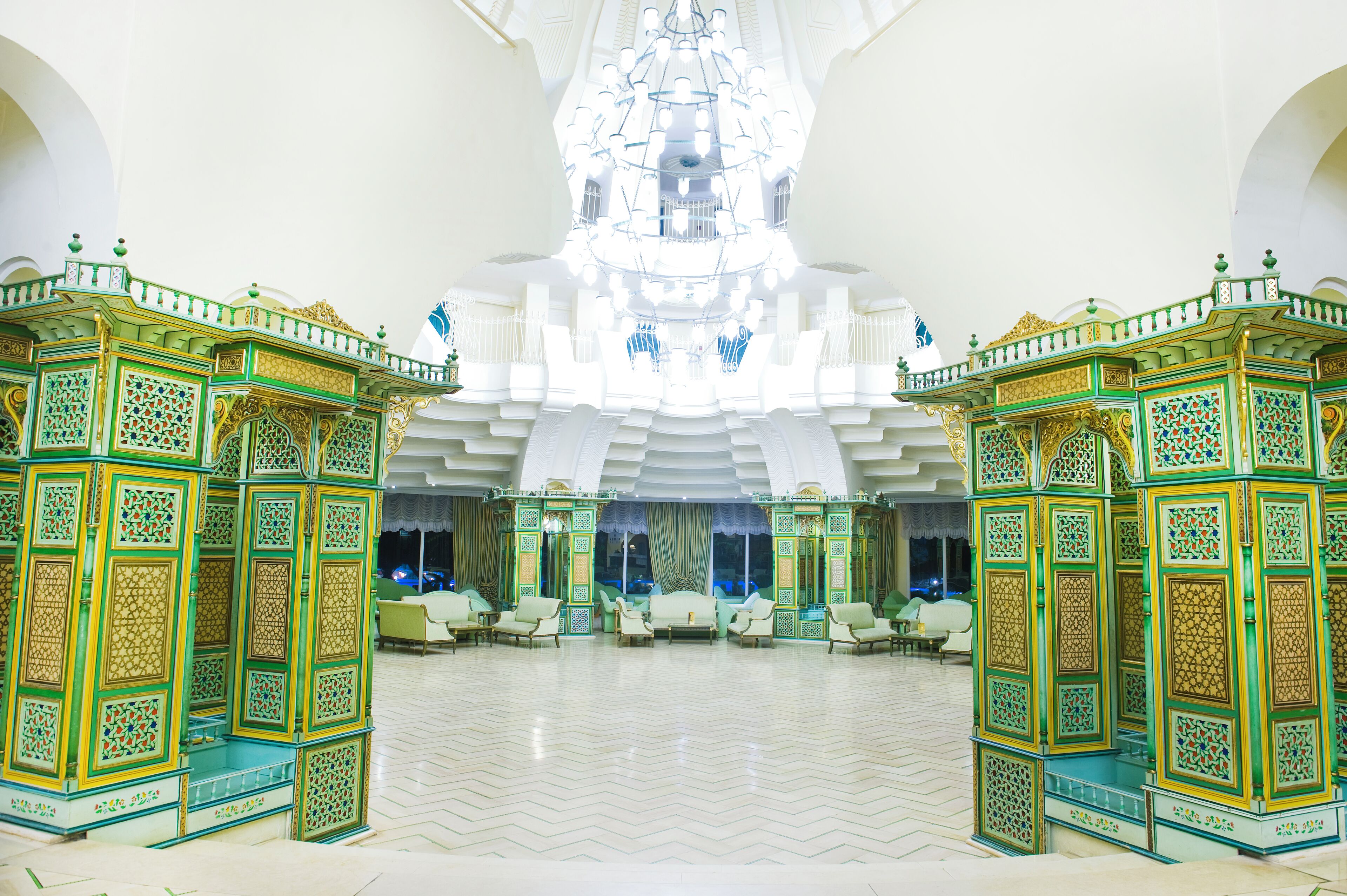 A spacious lobby with ornate green and gold decor, a large chandelier, and a seating area with sofas and chairs.