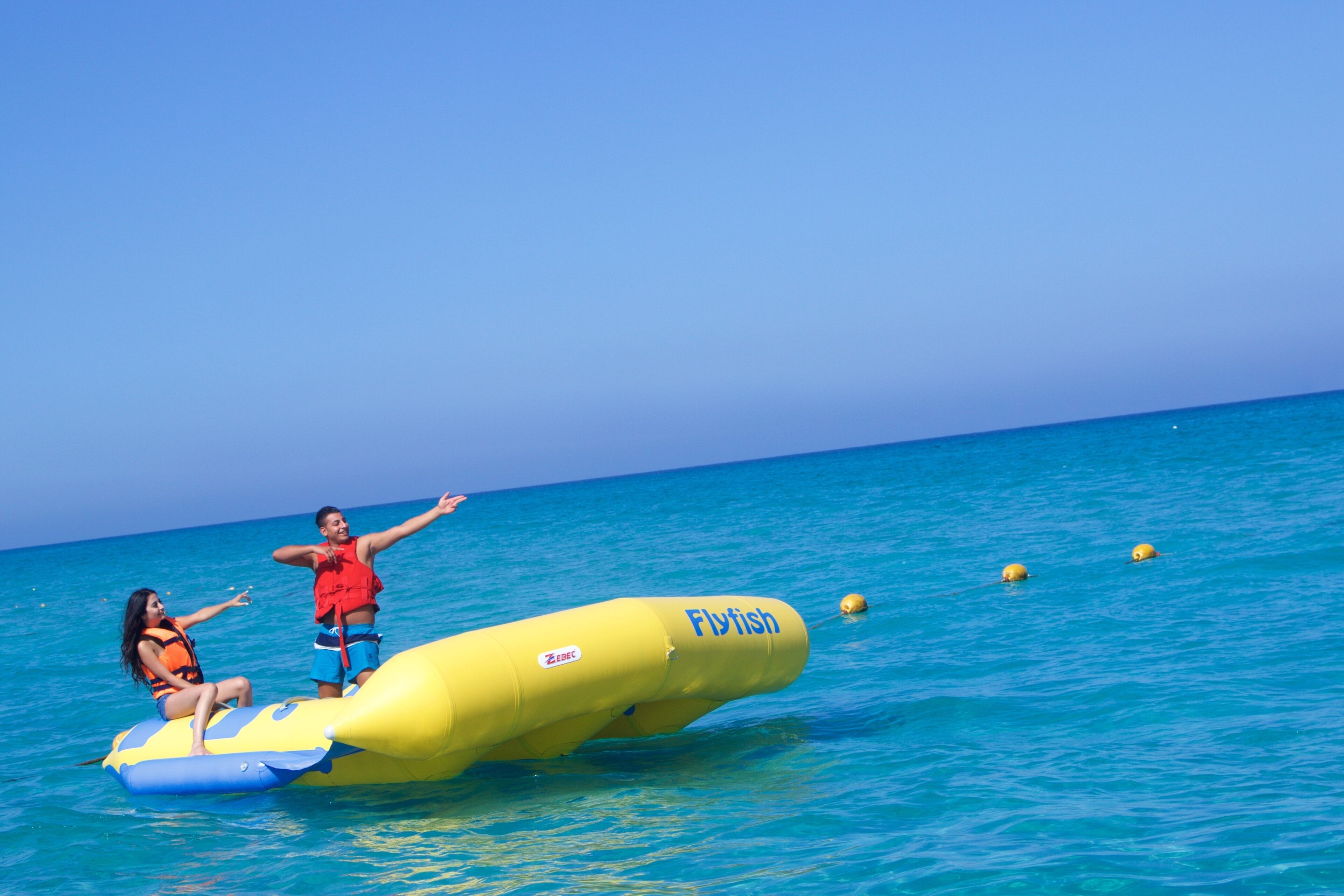 Two people on a yellow inflatable raft labeled 'Flyfish' in a clear blue sea.
