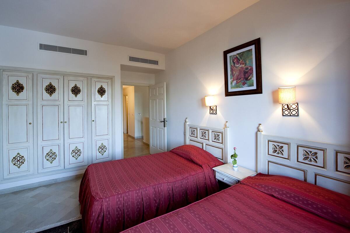 A hotel room with two beds, a white wardrobe, a painting on the wall, and a nightstand with a lamp.