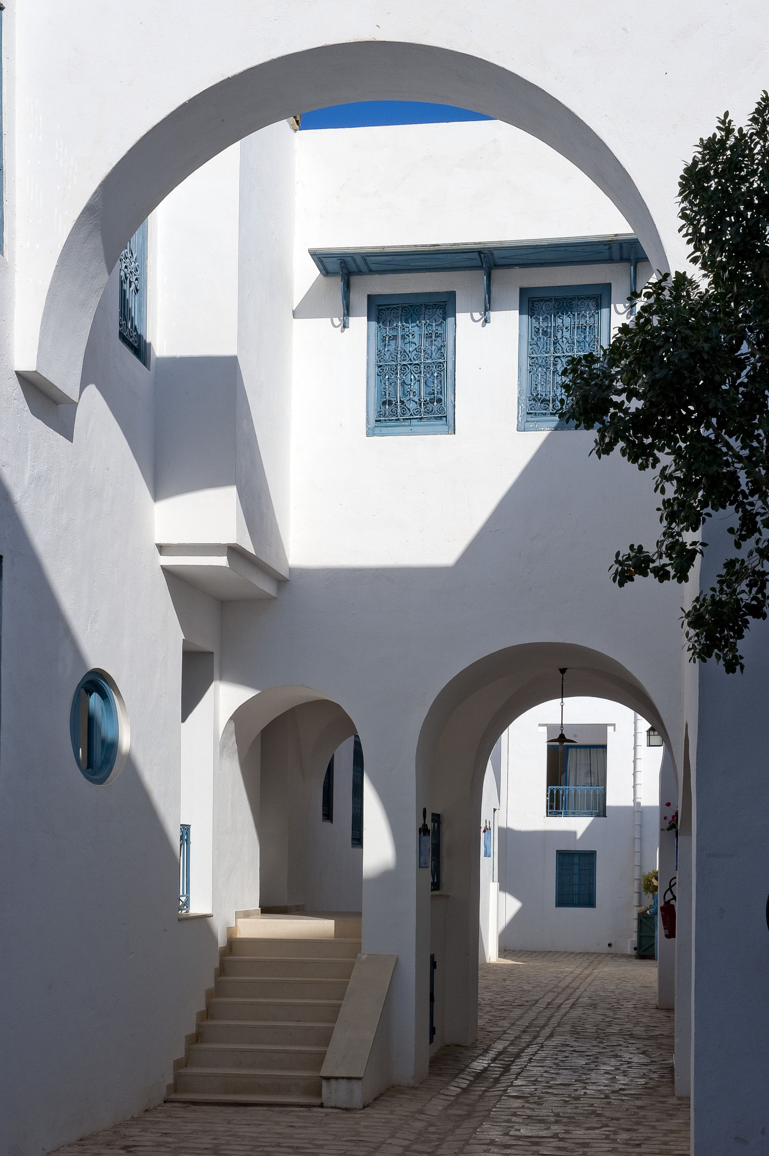 Un estrecho callejón adoquinado con paredes blancas y marcos de ventanas azules, que conduce a un edificio con una escalera.