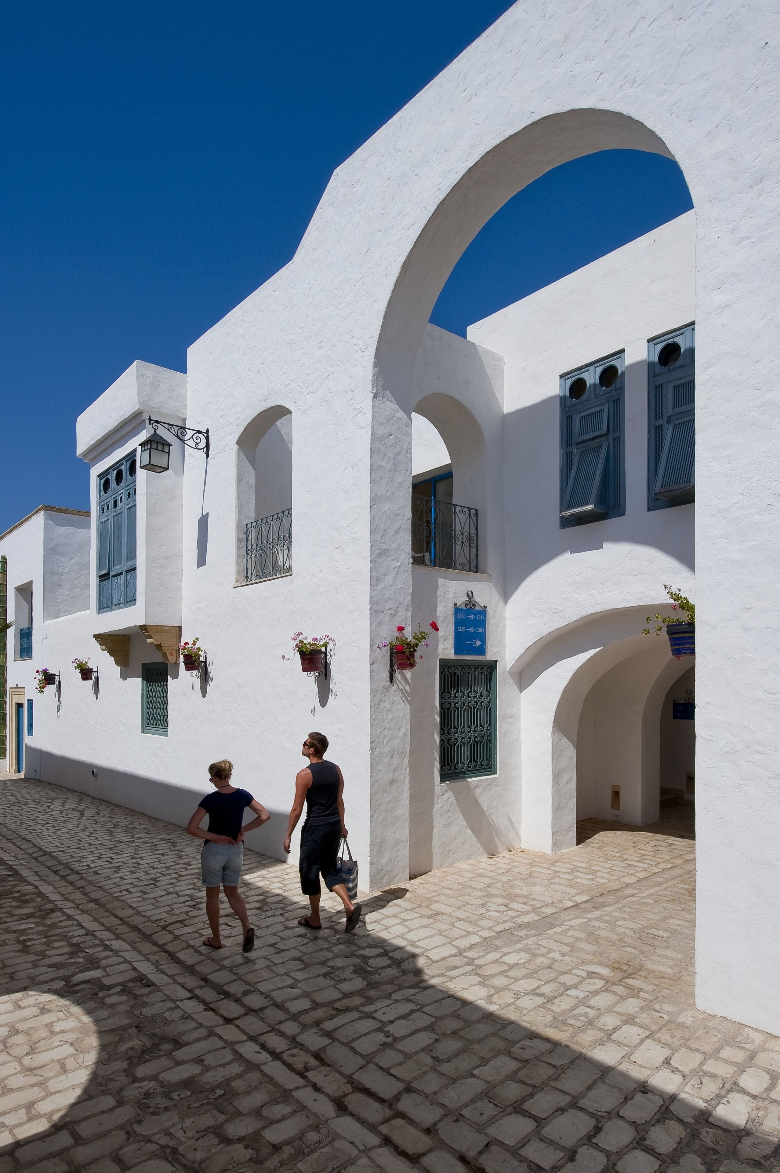 A white building with arched doorways and windows, a cobblestone street, and hanging flower baskets.