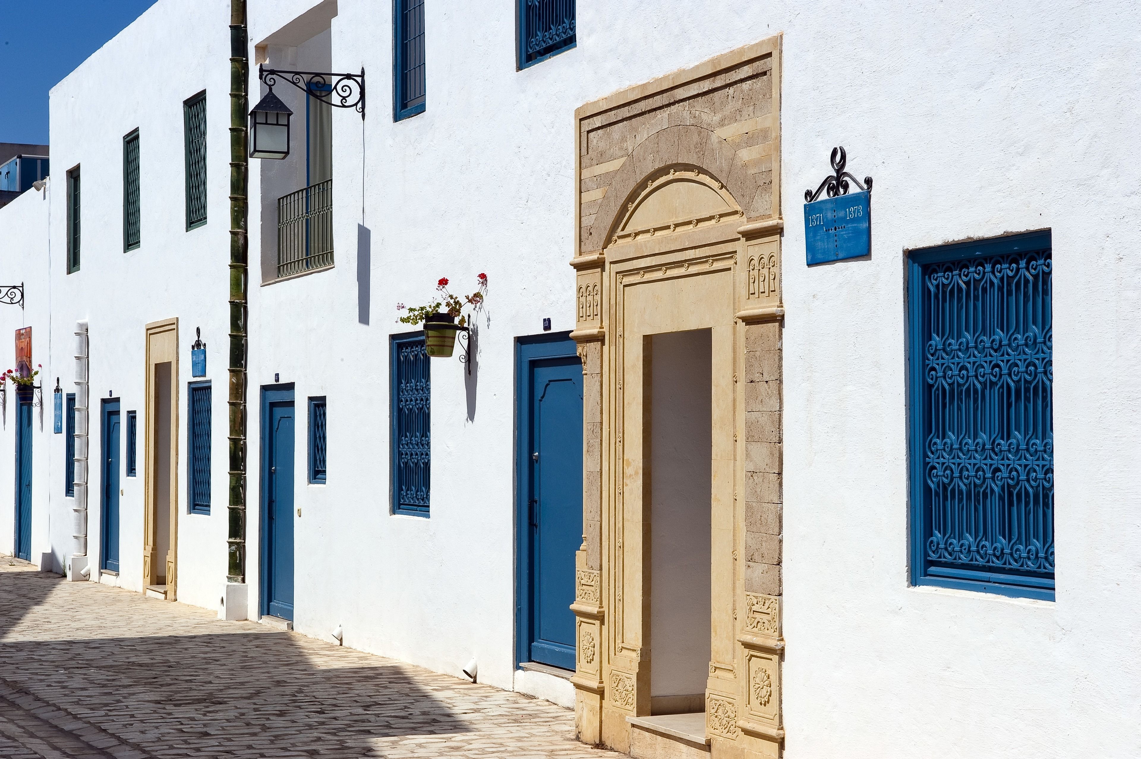 A row of white buildings with blue doors and windows, a cobblestone street, and a clear blue sky.