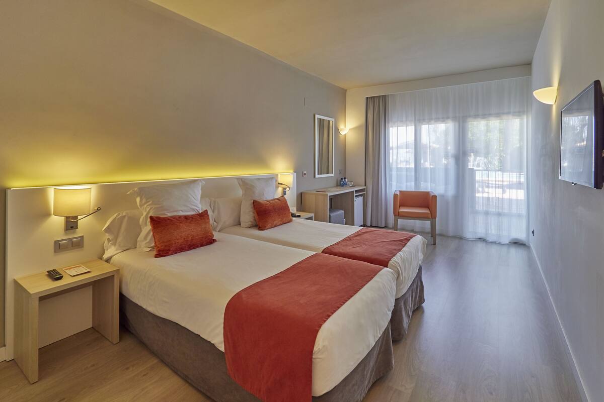 Camera d'albergo con due letti, una scrivania, una sedia e un'ampia finestra con tende.
