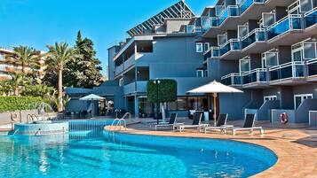 Une piscine entourée de chaises longues et de parasols, bordée d’un immeuble à plusieurs étages doté de balcons, le tout sous un ciel d’un bleu limpide.