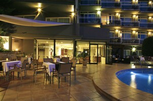Un espacio exterior para comer con mesas y sillas, una piscina y un edificio con varios balcones.