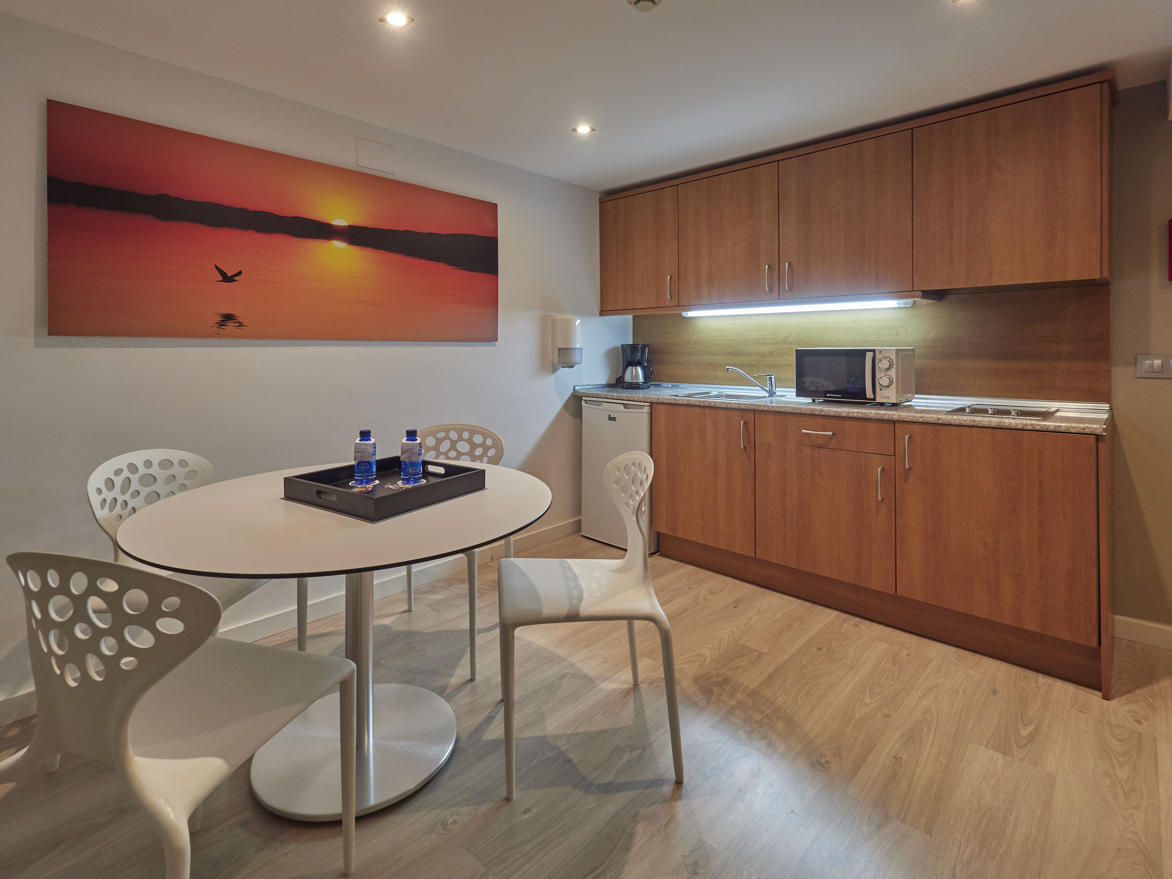 Una cocina moderna con armarios de madera, microondas y cafetera. Hay una mesa de comedor con sillas y un cuadro de un atardecer con aves.