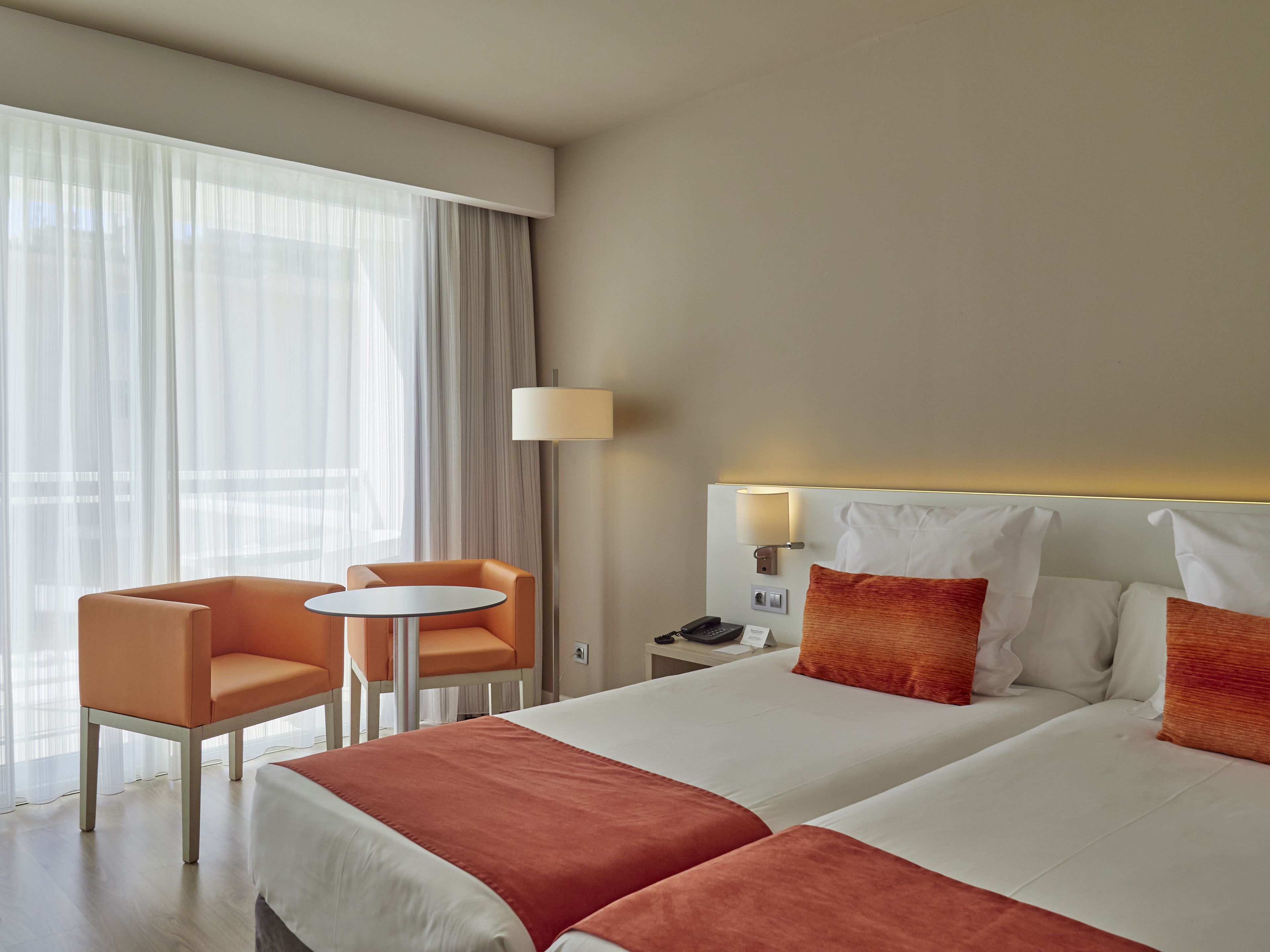 A hotel room with two beds, a small table, two chairs, a lamp, and a nightstand with a phone and a sign.