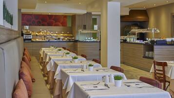 Een restaurant met lange tafels klaargezet voor diners, een keuken op de achtergrond en een modern interieur.