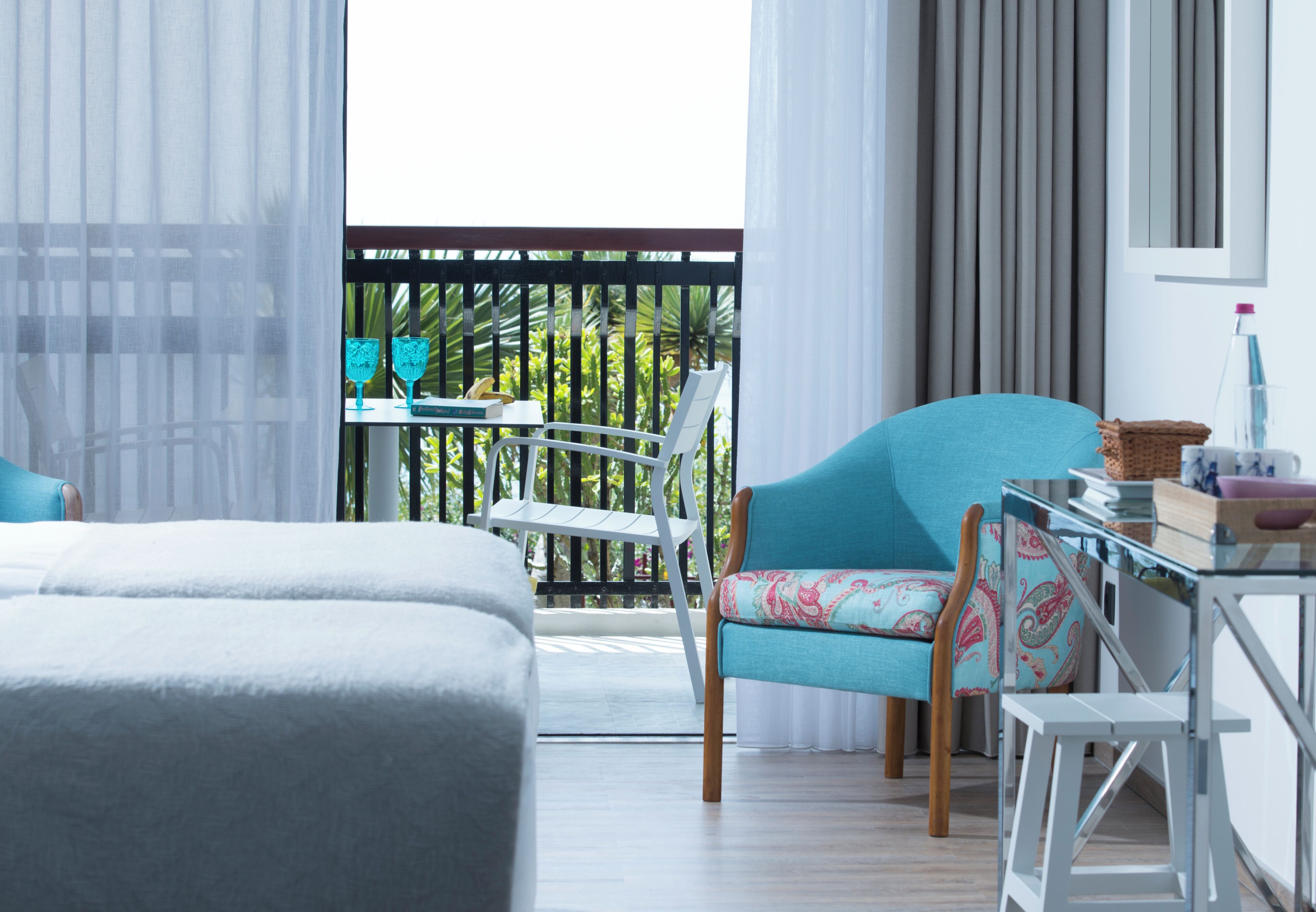 A hotel room with a balcony, a bed, a chair, a table, and a window with curtains.