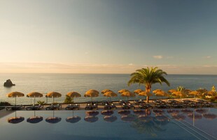 An infinity pool with lounge chairs and umbrellas, overlooking the ocean with a palm tree and a rock formation in the distance.