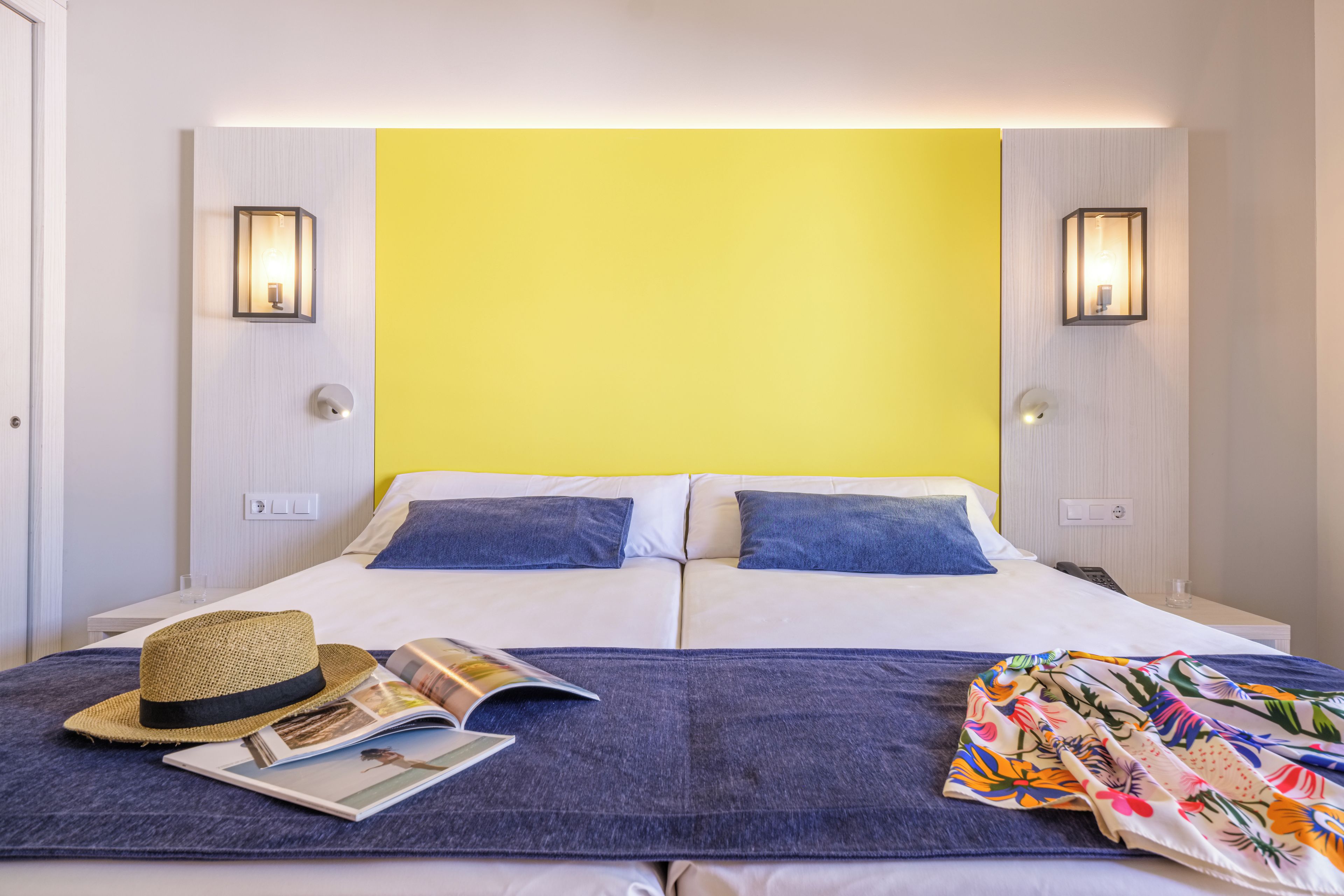 Una cama doble con dos almohadas, un sombrero, una revista y un textil colorido sobre la colcha.