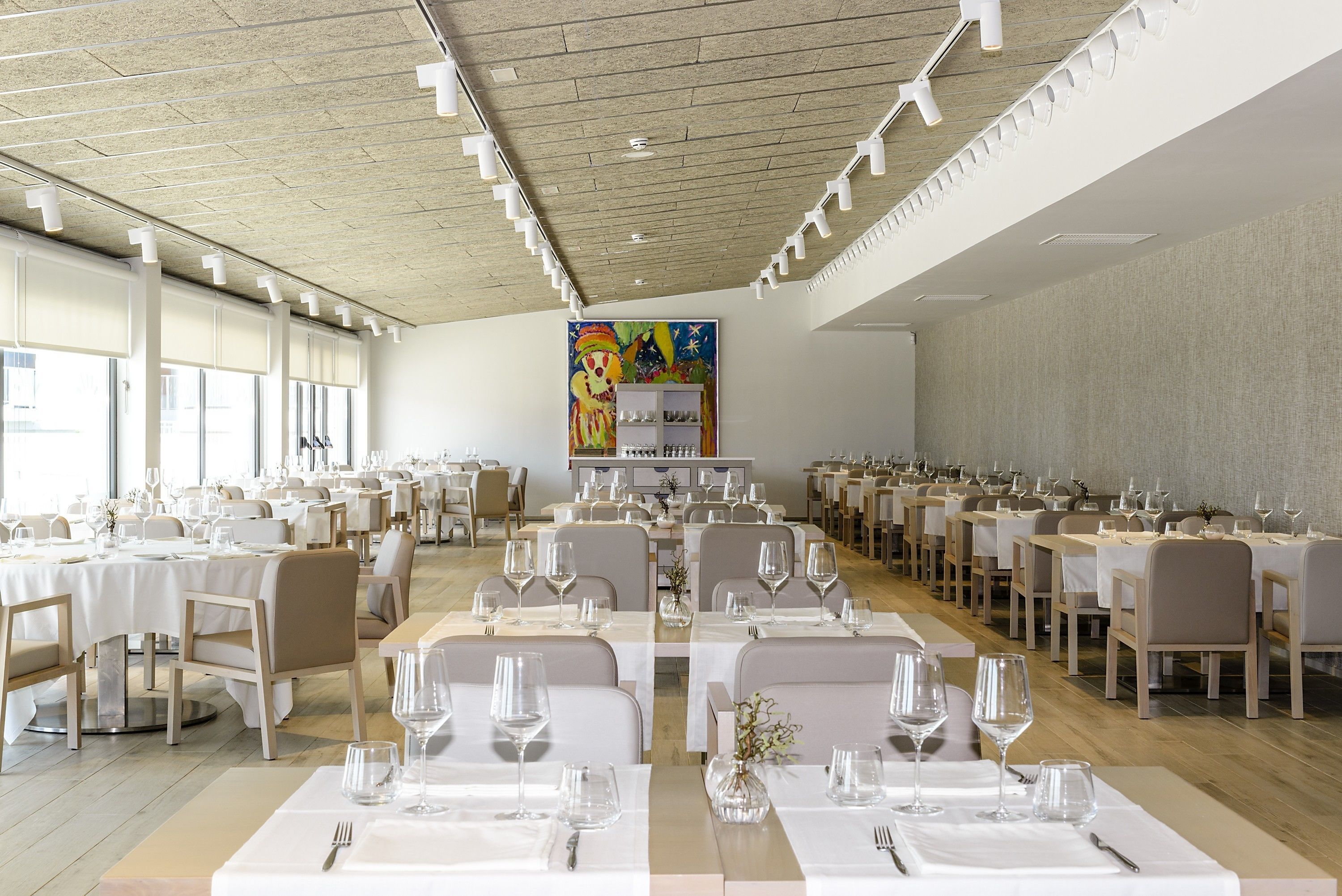 Un amplio comedor con varias mesas dispuestas para el servicio, grandes ventanales en un lateral y un colorido mural en la pared de fondo.