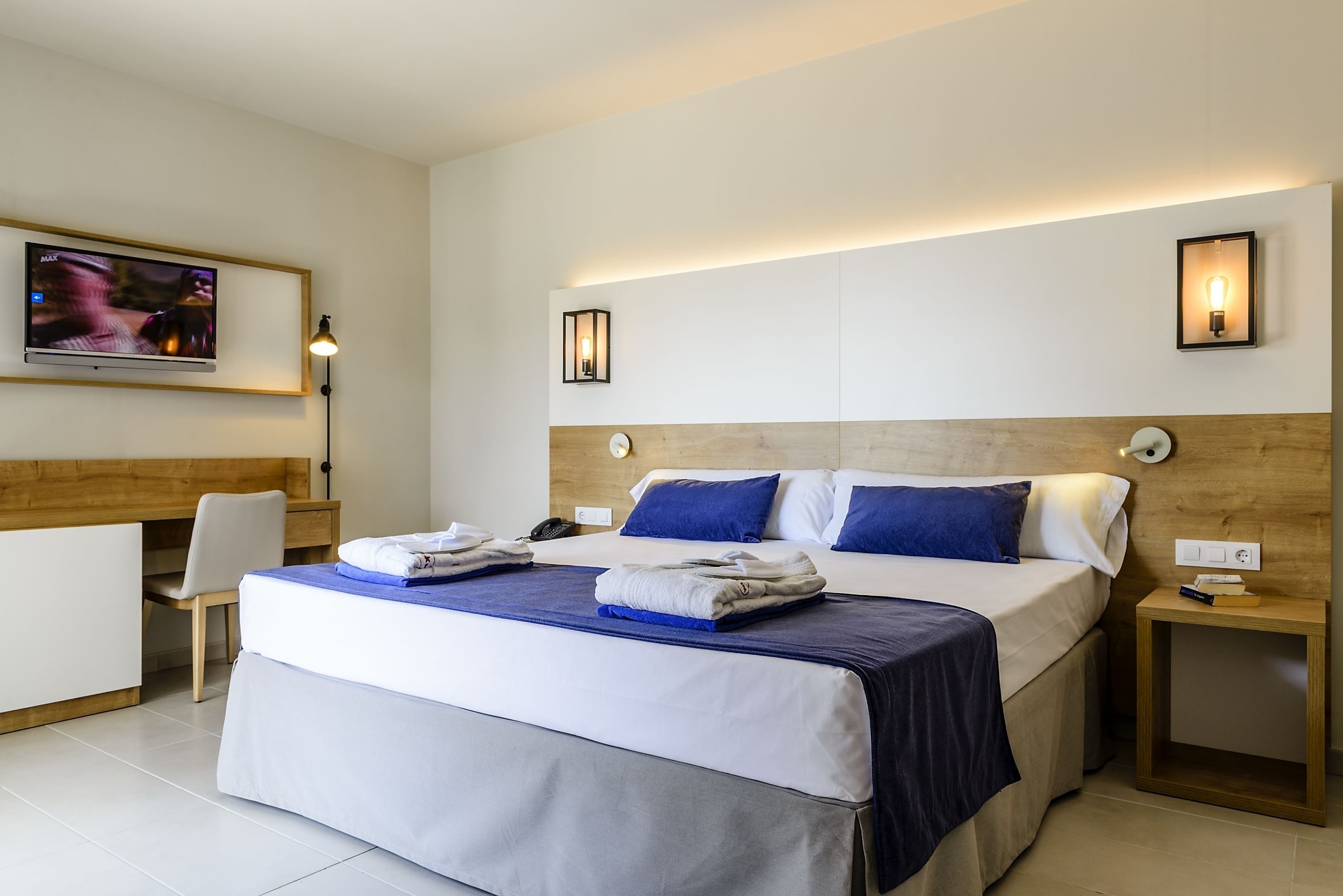 A hotel room with a large bed, a desk, a chair, a television, and two wall-mounted lights.