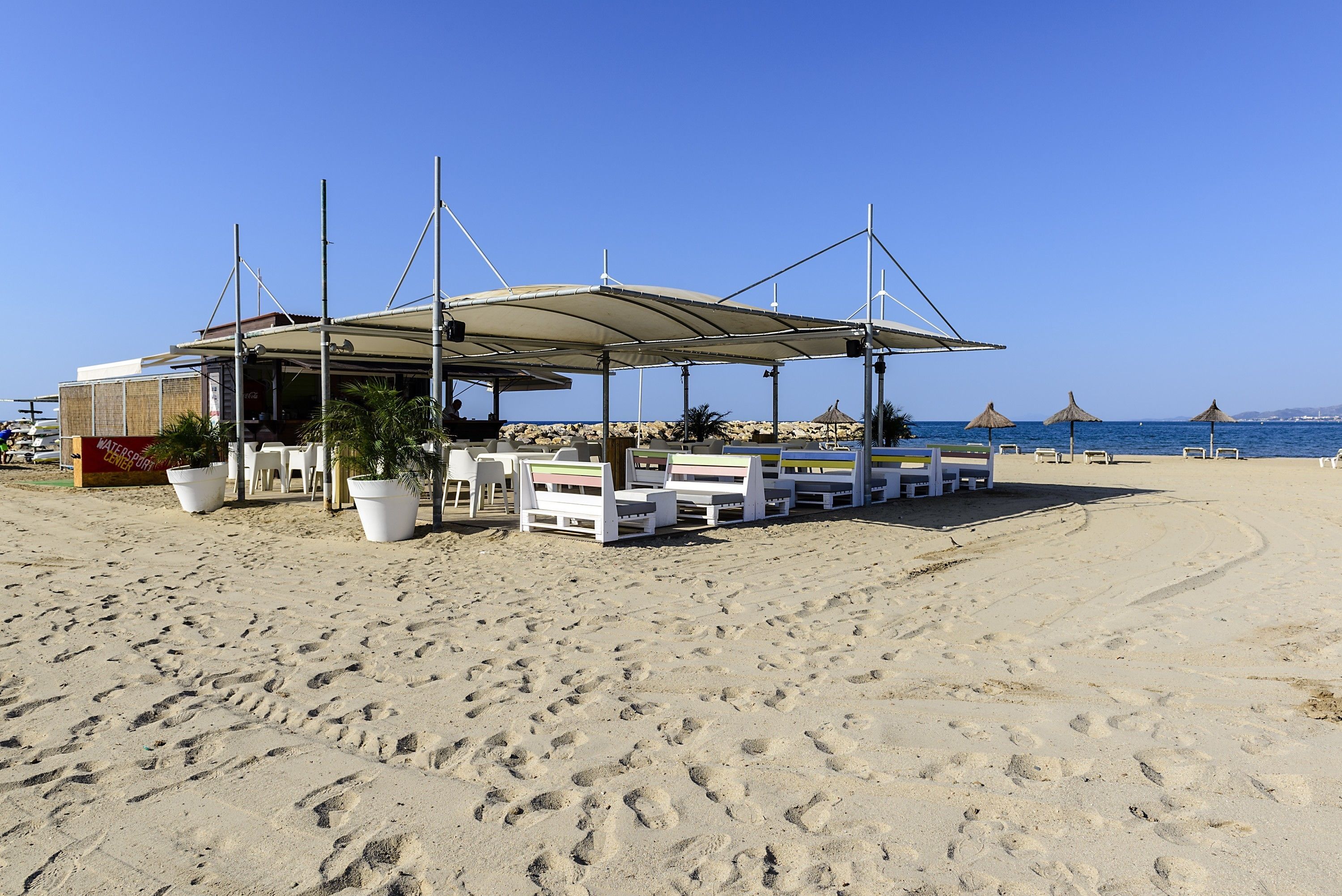 Un café frente a la playa con mesas al aire libre y un área techada, con vistas al mar.